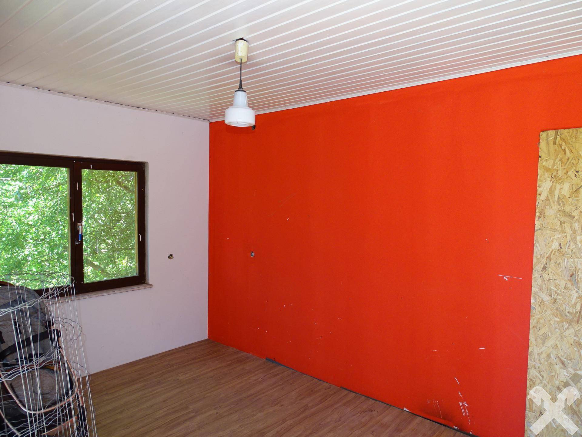 Leerer Raum mit leuchtend oranger Wand, Holzfußboden und Blick ins Grüne durch das Fenster.