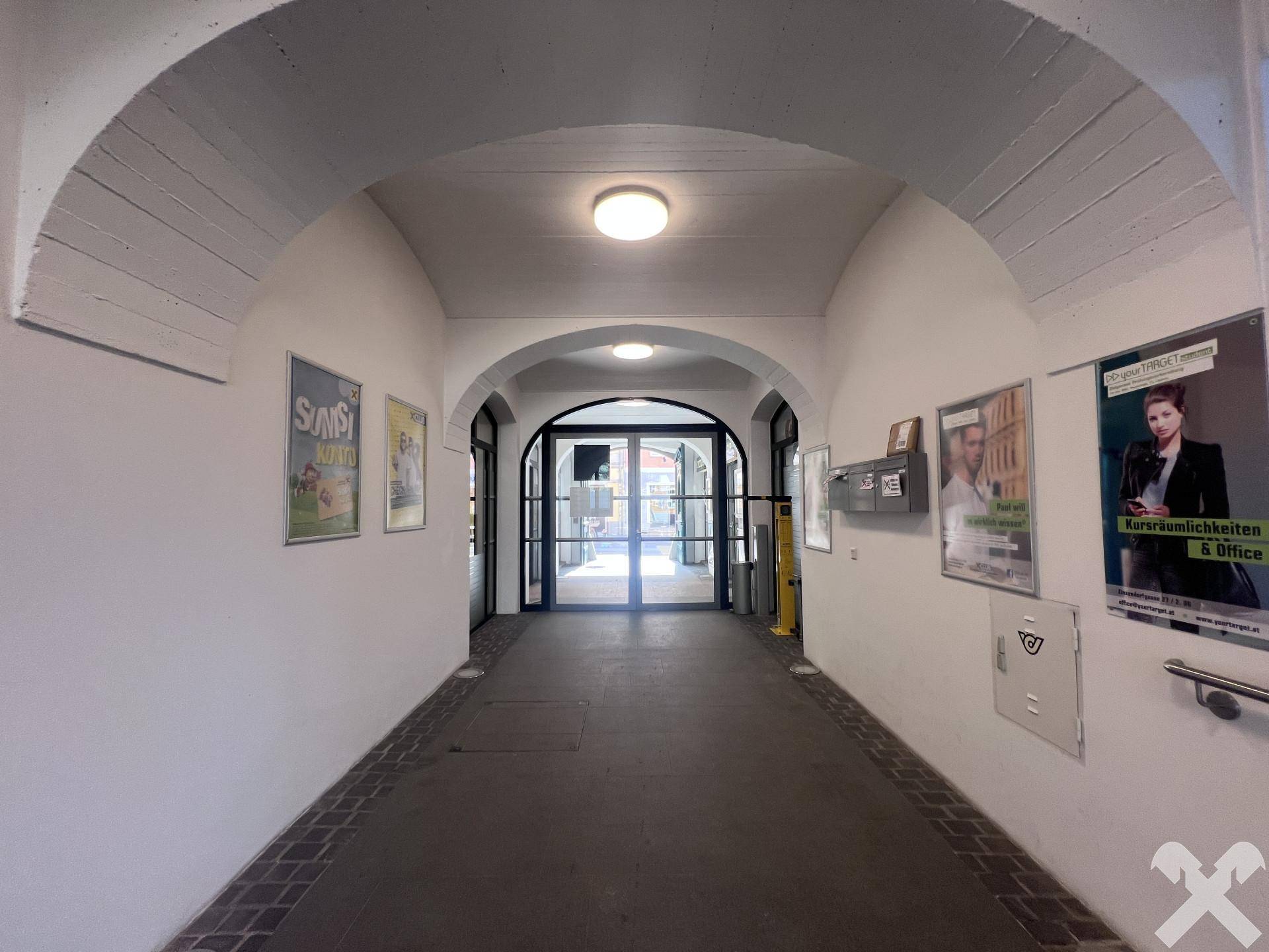 Langer Flur mit gewölbten Decken und Plakaten an den Wänden, der zu einem weiteren Bereich führt.