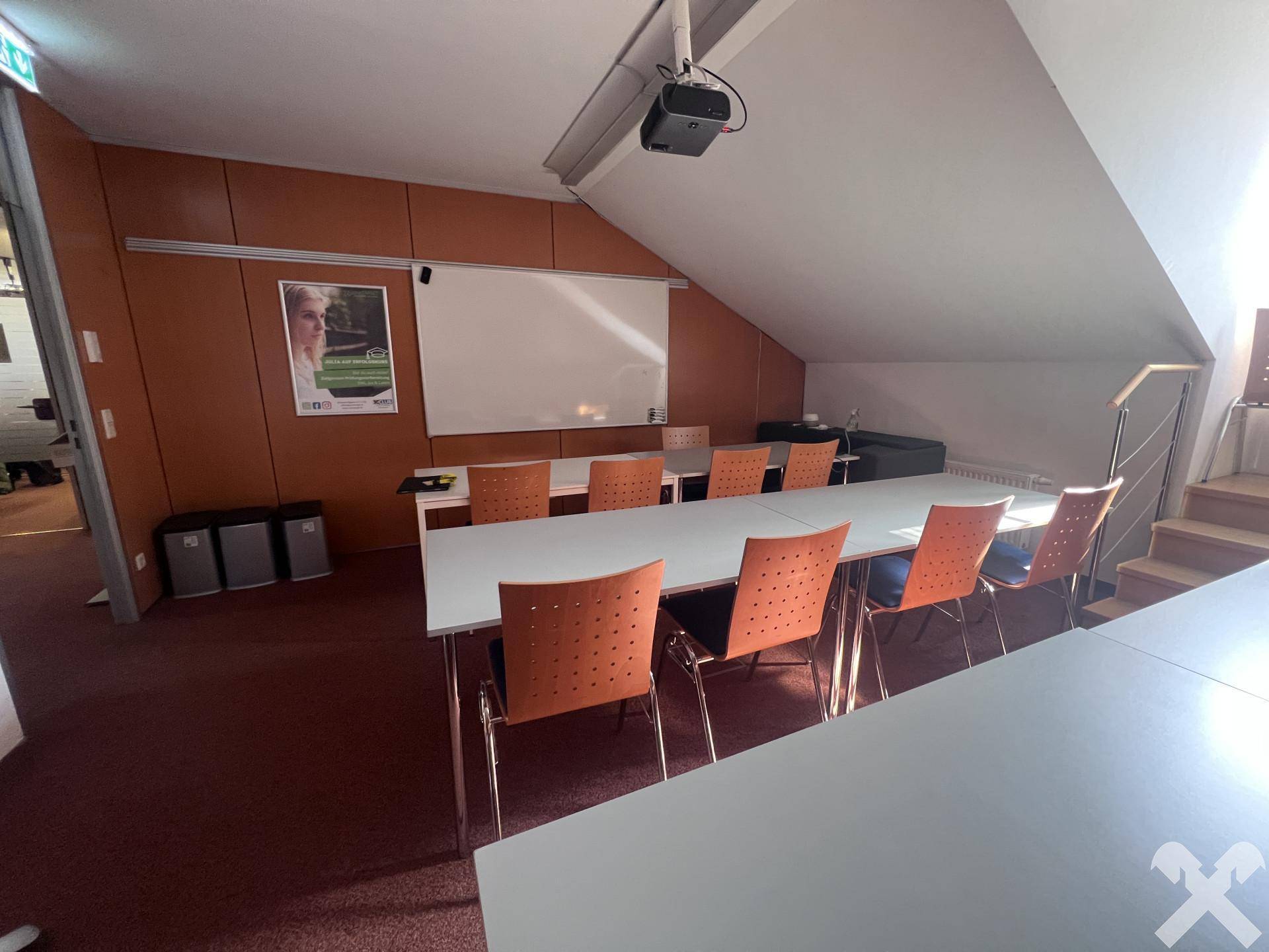 Besprechungsraum mit schräger Decke, Projektor und einem langen Tisch mit Stühlen.