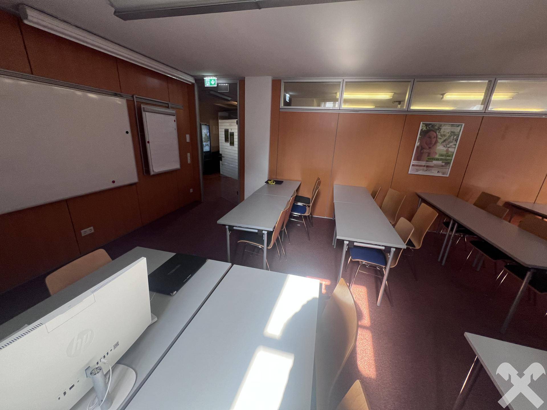 Schulungsraum mit mehreren Tischen und Stühlen, sowie Fenstern im oberen Bereich der Wand.