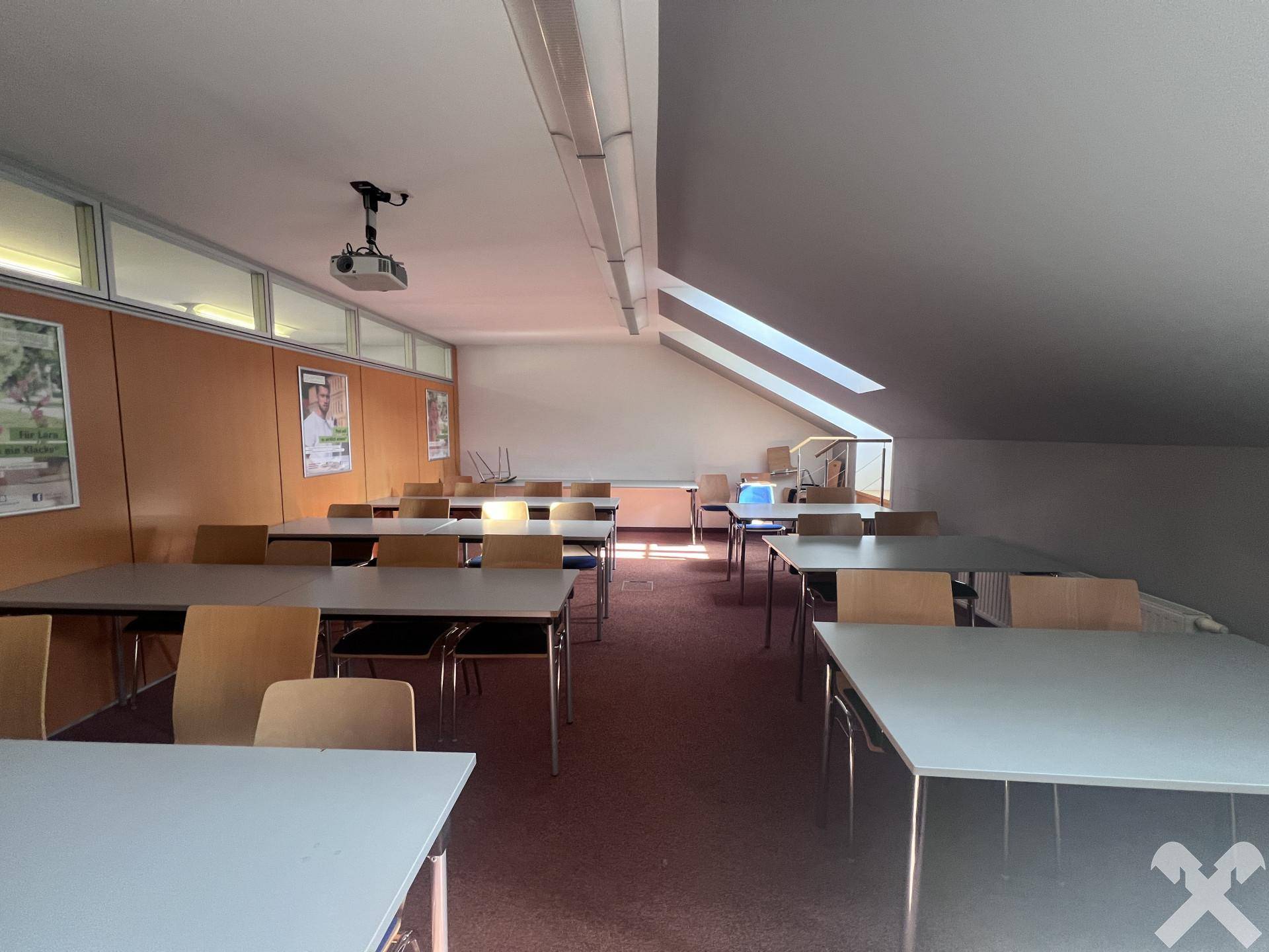 Großer Schulungsraum mit schräger Decke, Dachfenstern und vielen Tischen für Lernzwecke.
