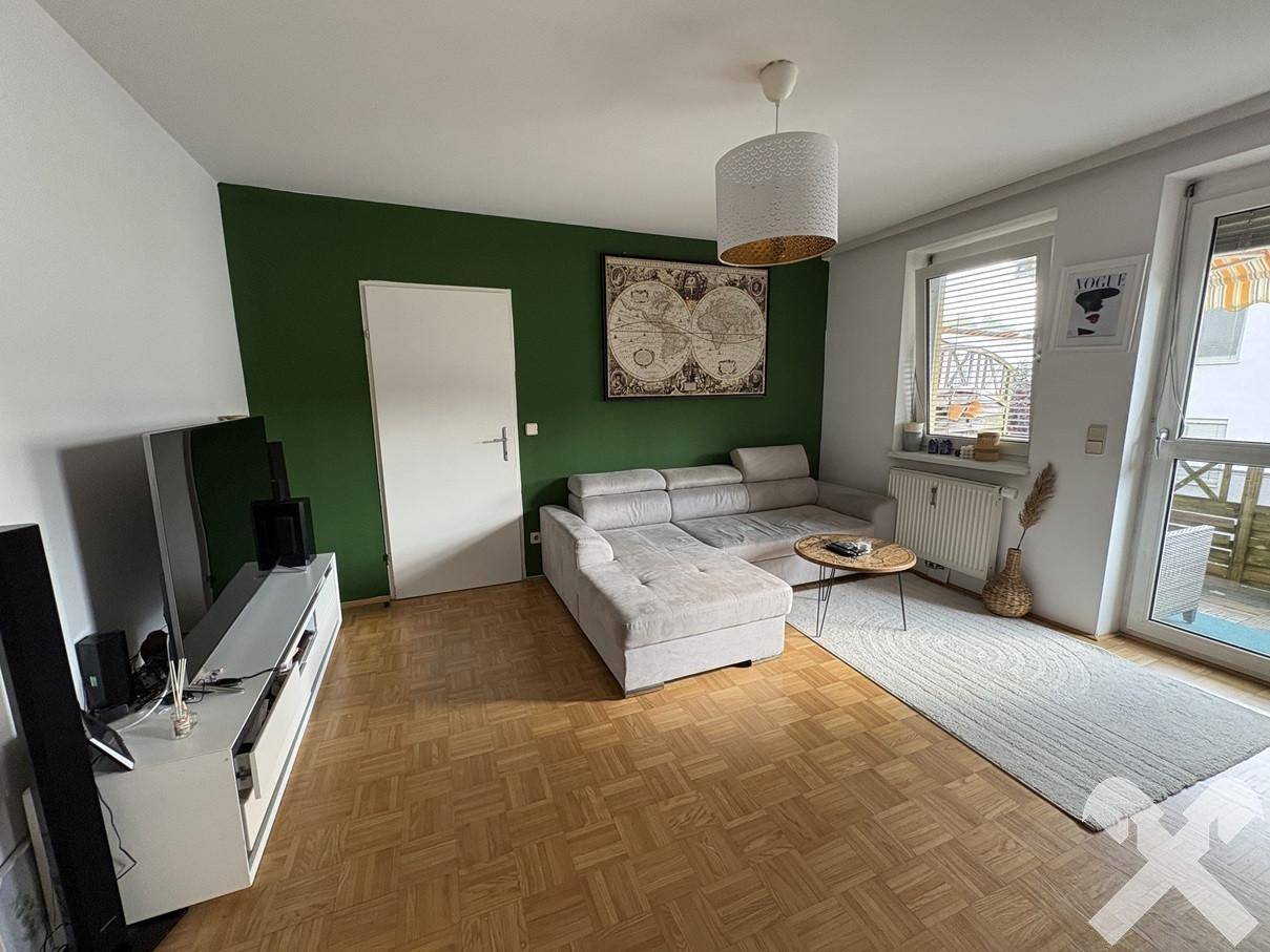 Helles Wohnzimmer mit Sofa, Fernseher und Zugang zu einem Balkon, mit viel Tageslicht.