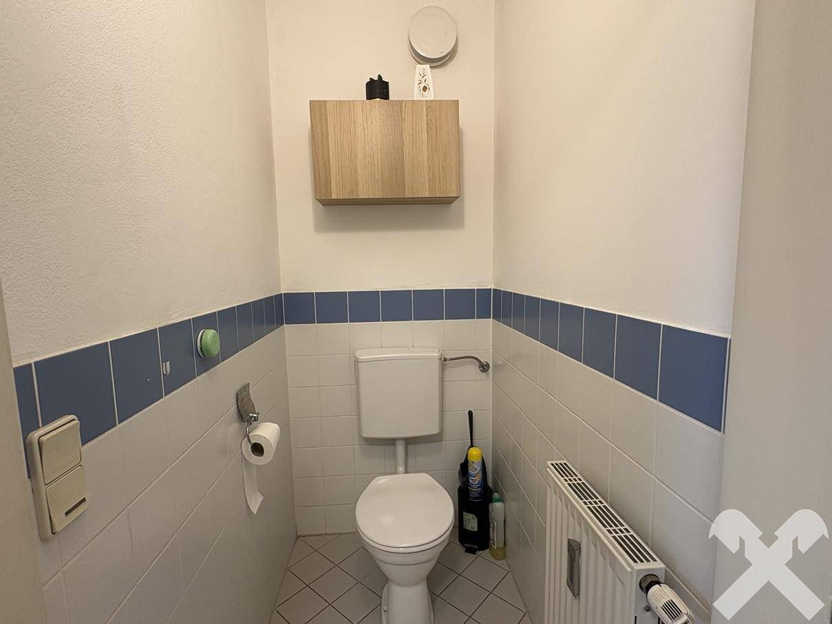 Separates WC mit Toilette, Heizkörper und Wandregal, gefliest in Weiß und Blau.