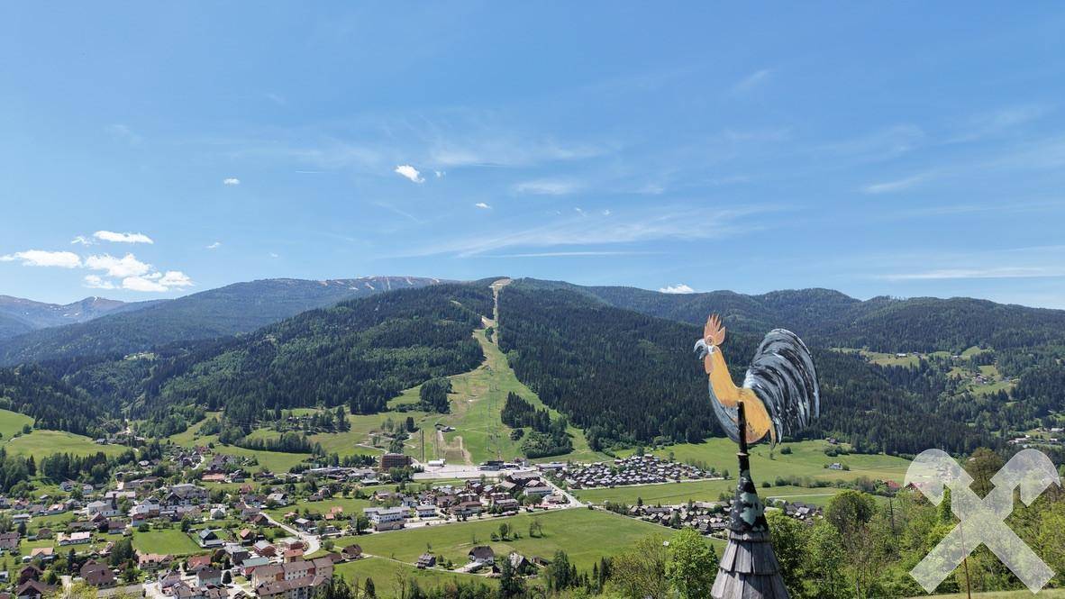 Panoramablick über ein malerisches Tal mit einem Dorf und bewaldeten Bergen unter blauem Himmel.