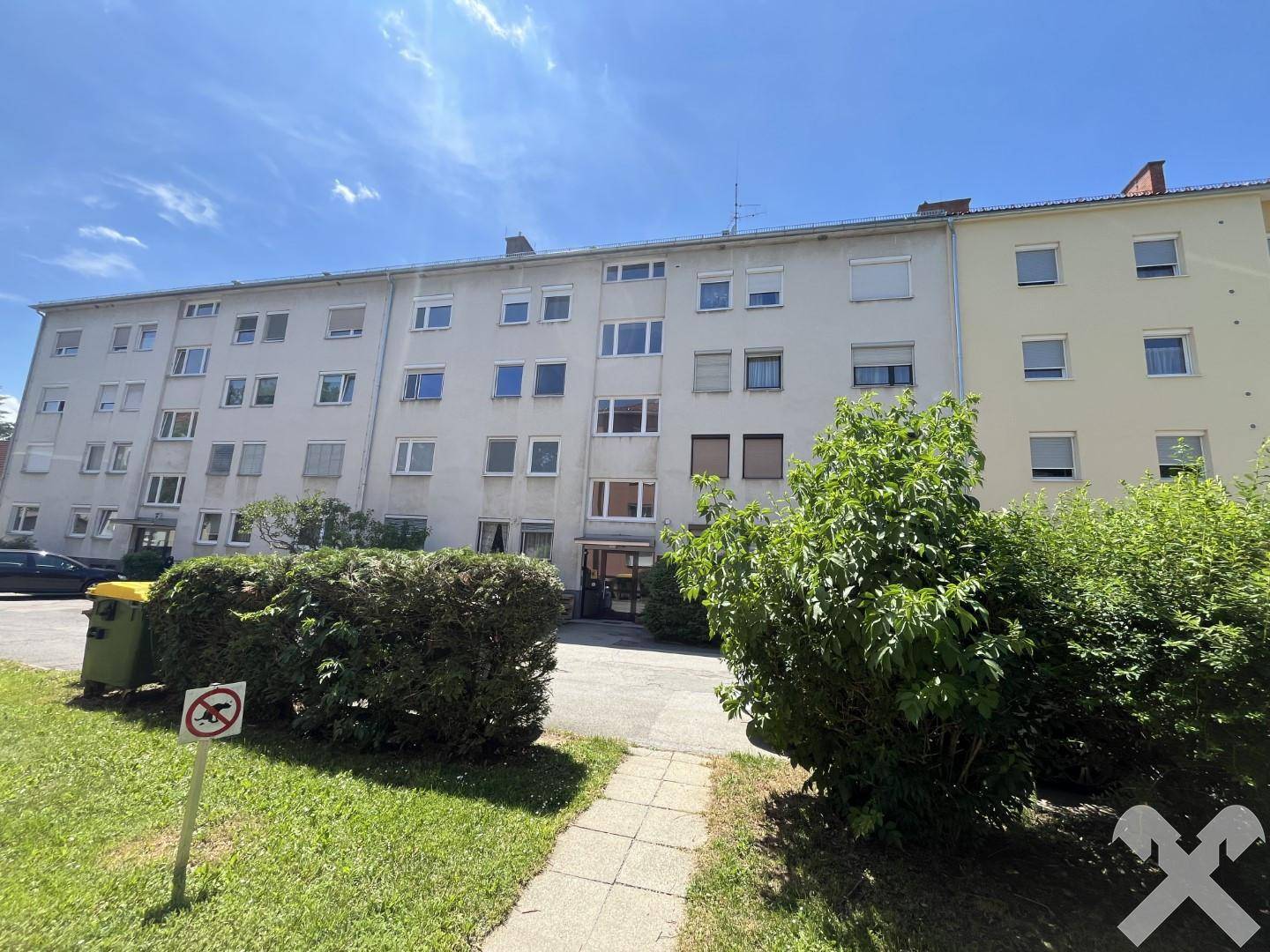 Ansicht der Gebäudefassade eines Mehrfamilienhauses mit Eingangsbereich, gepflegtem Rasen und Büschen.