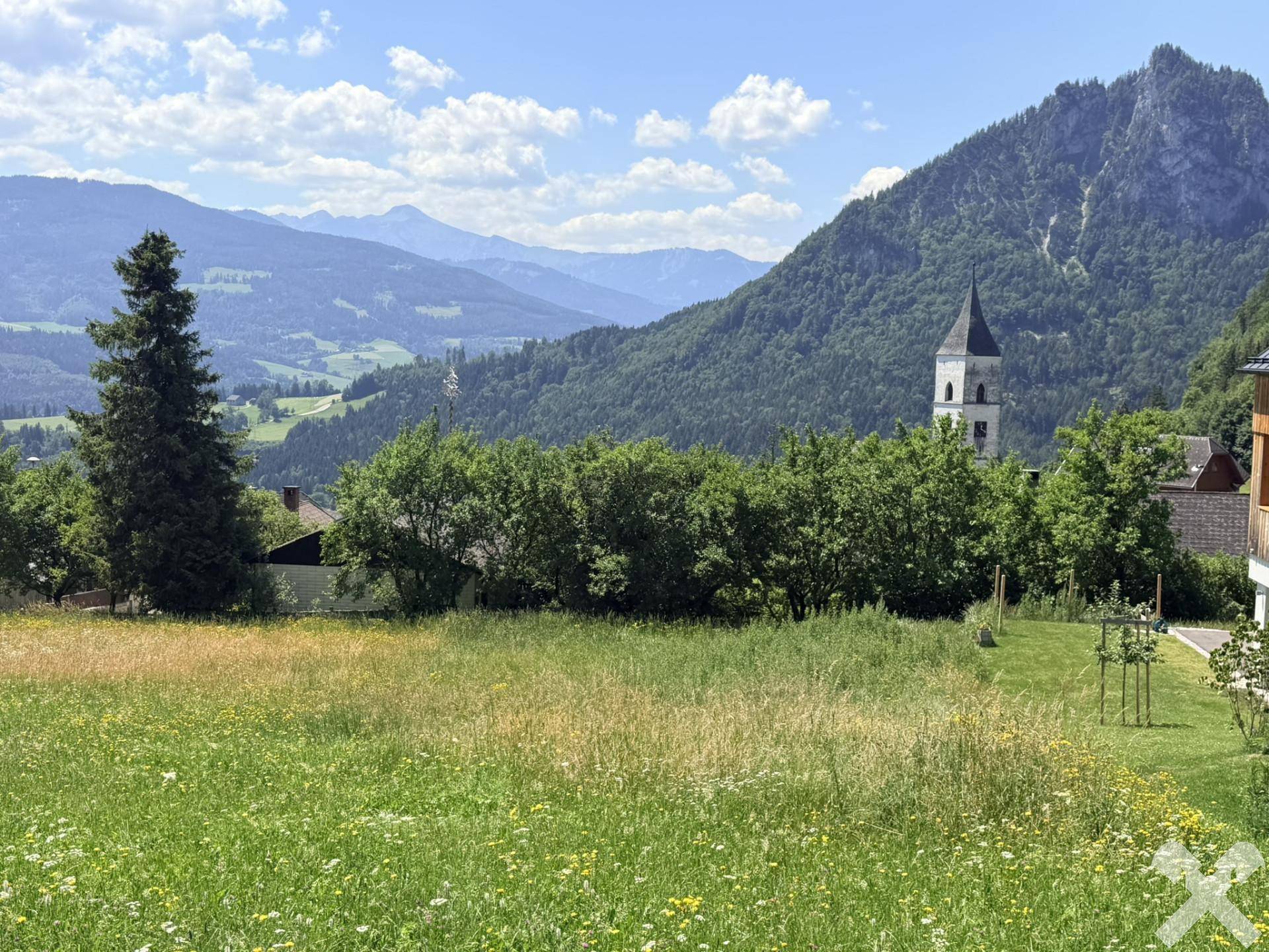Weitläufige grüne Wiesen mit Blick auf die umliegende Berglandschaft und einen Kirchturm.