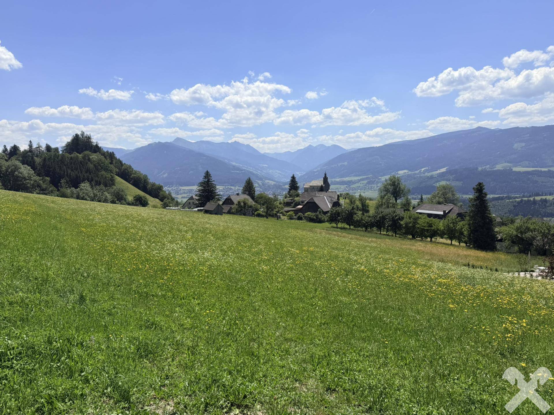 Ausgedehnte grüne Hügellandschaft mit einem Dorf und fernen Bergen unter einem strahlend blauen Himmel.