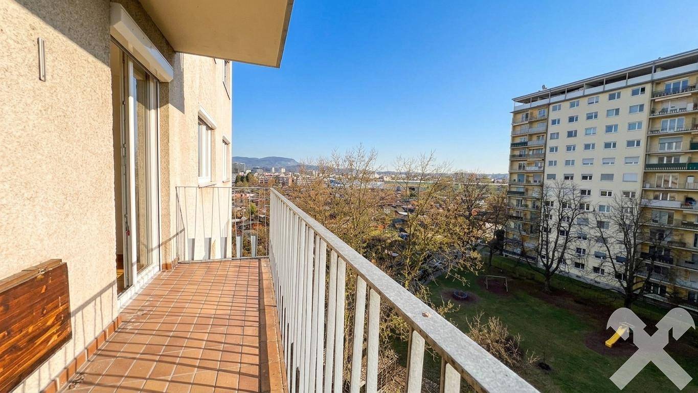 Geräumiger Balkon mit Fliesenboden, Sitzbank und Blick auf Bäume sowie umliegende Gebäude.