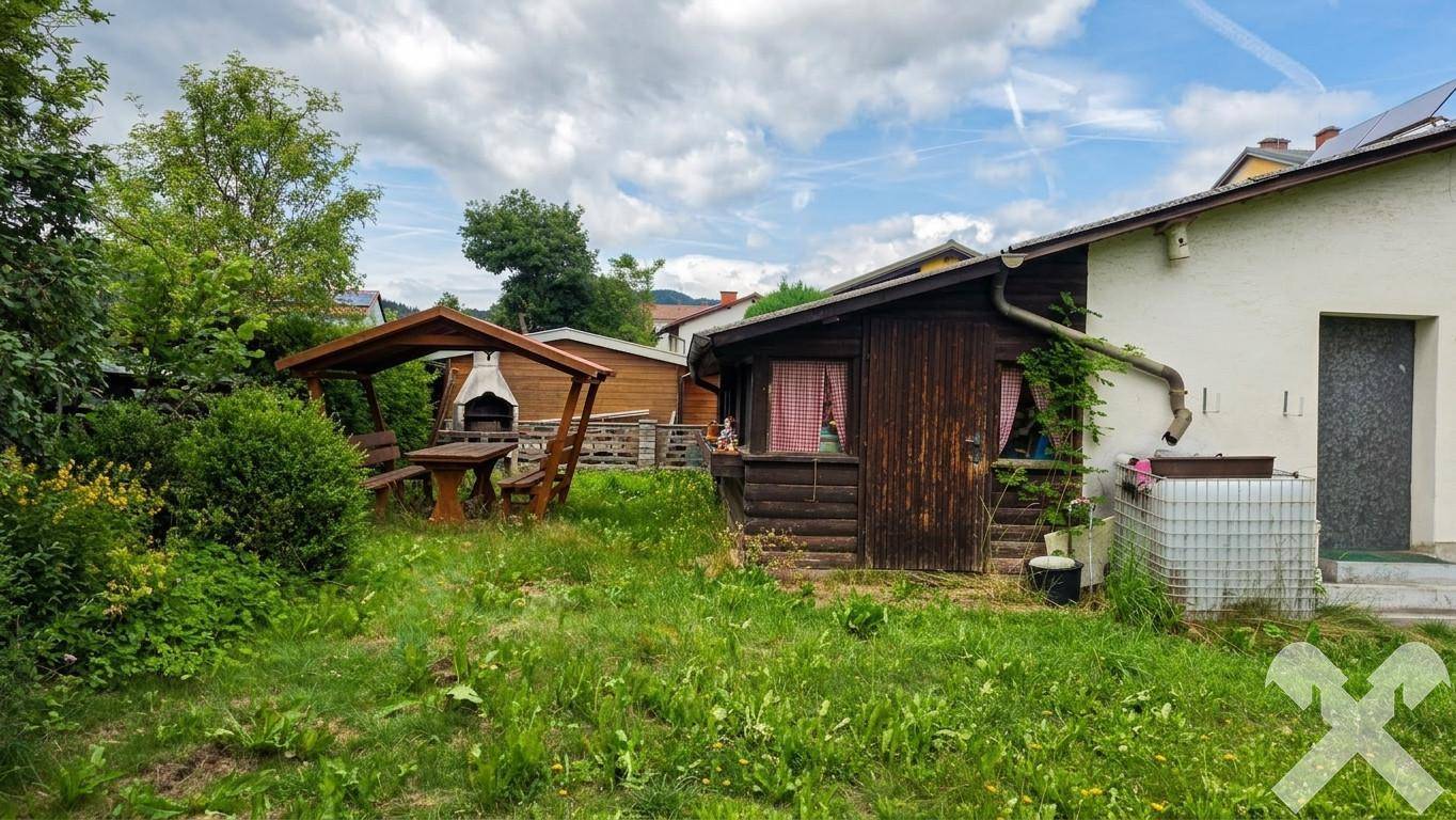 Gemütlicher Holzpavillon mit Picknicktisch und Grillbereich neben einem kleinen Holzschuppen im Garten.