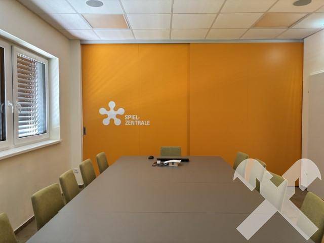 Konferenzraum mit langem Tisch, Stühlen und einer auffälligen orangefarbenen Wand mit Firmenlogo.