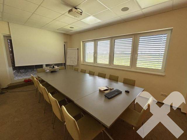 Konferenzraum mit langem Tisch, Stühlen, Beamer und Leinwand, ausgestattet mit Fenstern und Jalousien.