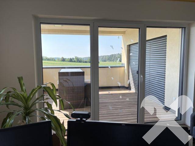 Blick aus einem Bürofenster auf eine Loggia mit Holzdeck und Gartenmöbeln, mit weitem Blick in die Landschaft.