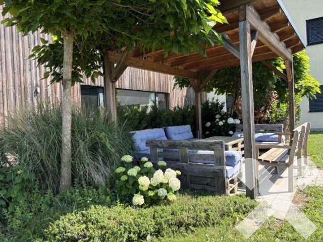 Gemütliche Holzpergola mit Palettenmöbeln und blauen Kissen in einem grünen Gartenbereich.