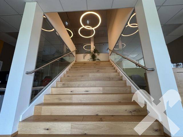 Moderne Treppe mit Holzstufen, Glasgeländer und Edelstahlhandläufen, beleuchtet von runden Deckenleuchten.