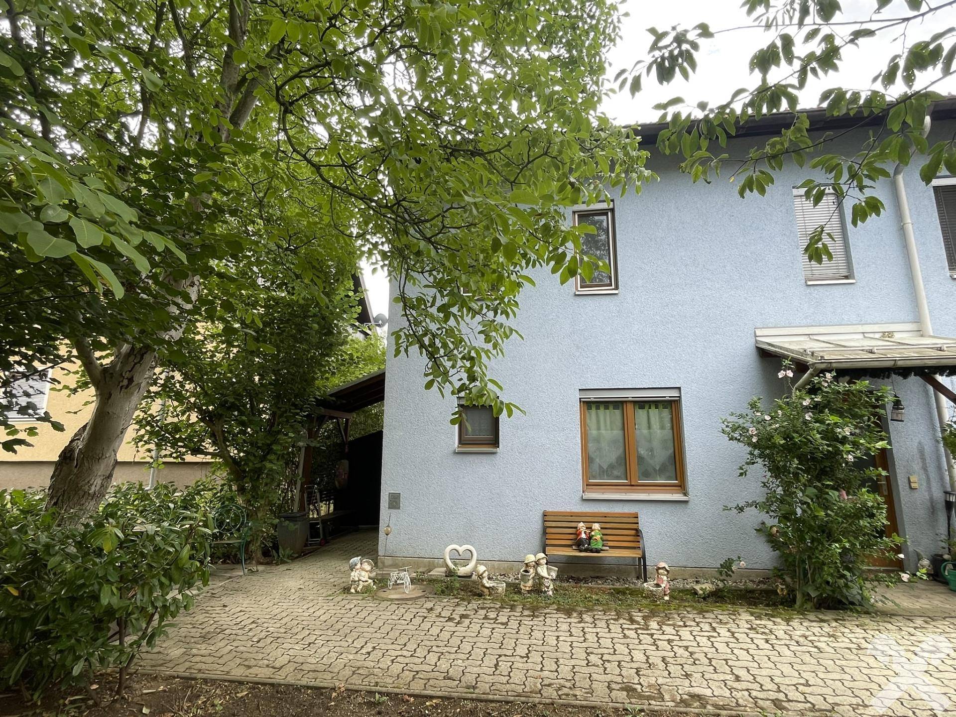 Seitlicher Eingangsbereich des Hauses mit gepflastertem Weg und einem kleinen Sitzbereich unter Bäumen.