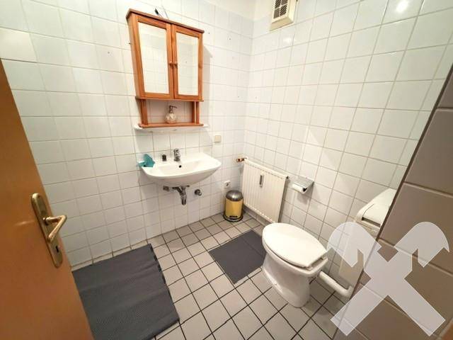 Badezimmer mit weißen Fliesen, Waschbecken, Toilette und einem Spiegelschrank.
