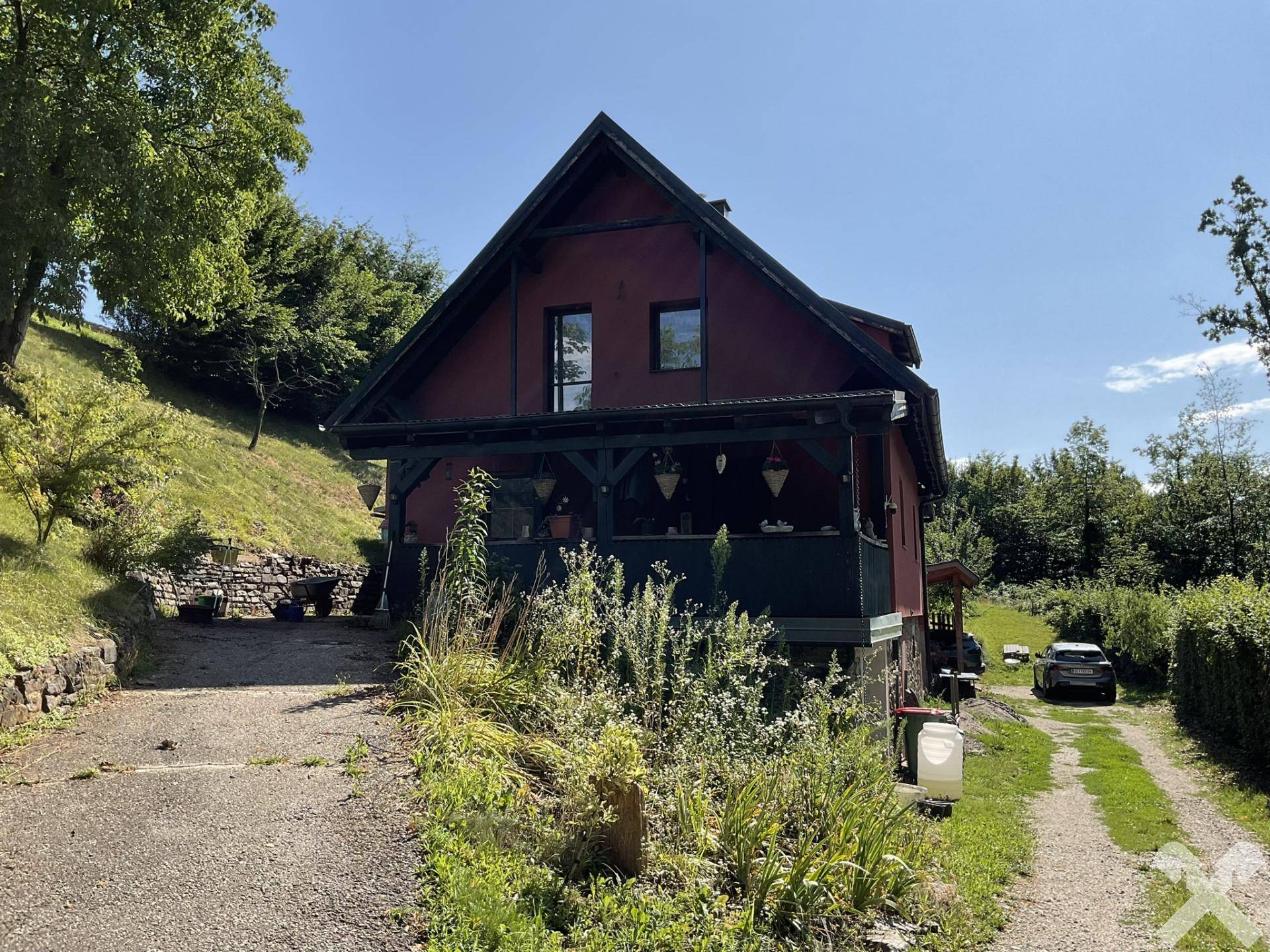 Vorderansicht des Hauses mit überdachtem Balkonbereich und einer Schotterauffahrt, umgeben von Grün.