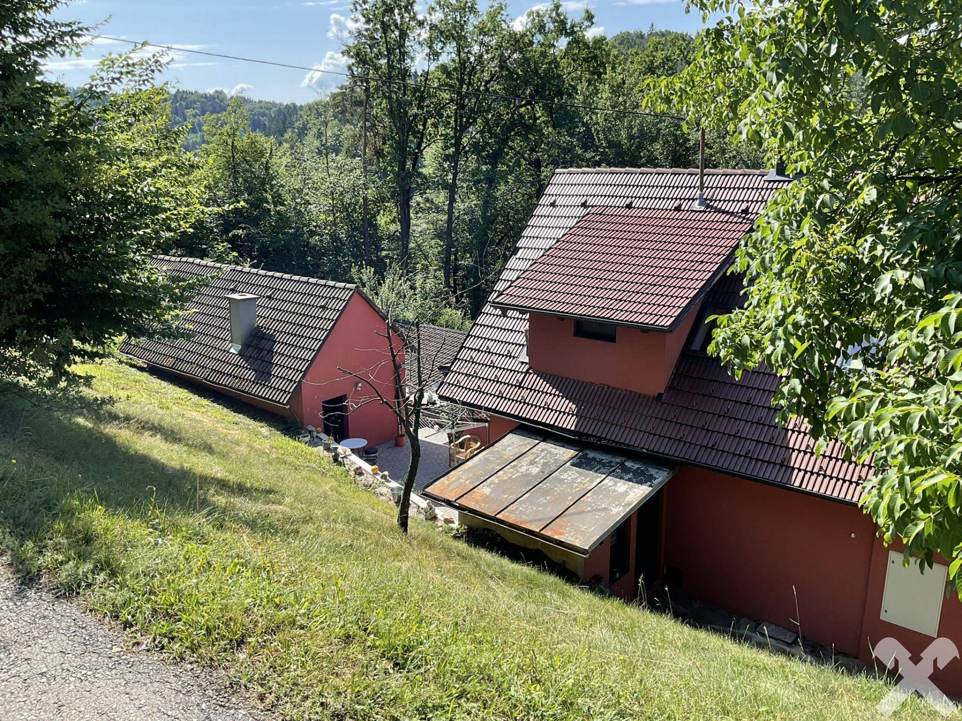 Ansicht des roten Hauses mit Nebengebäude und braunen Ziegeldächern, eingebettet in grüne Landschaft.