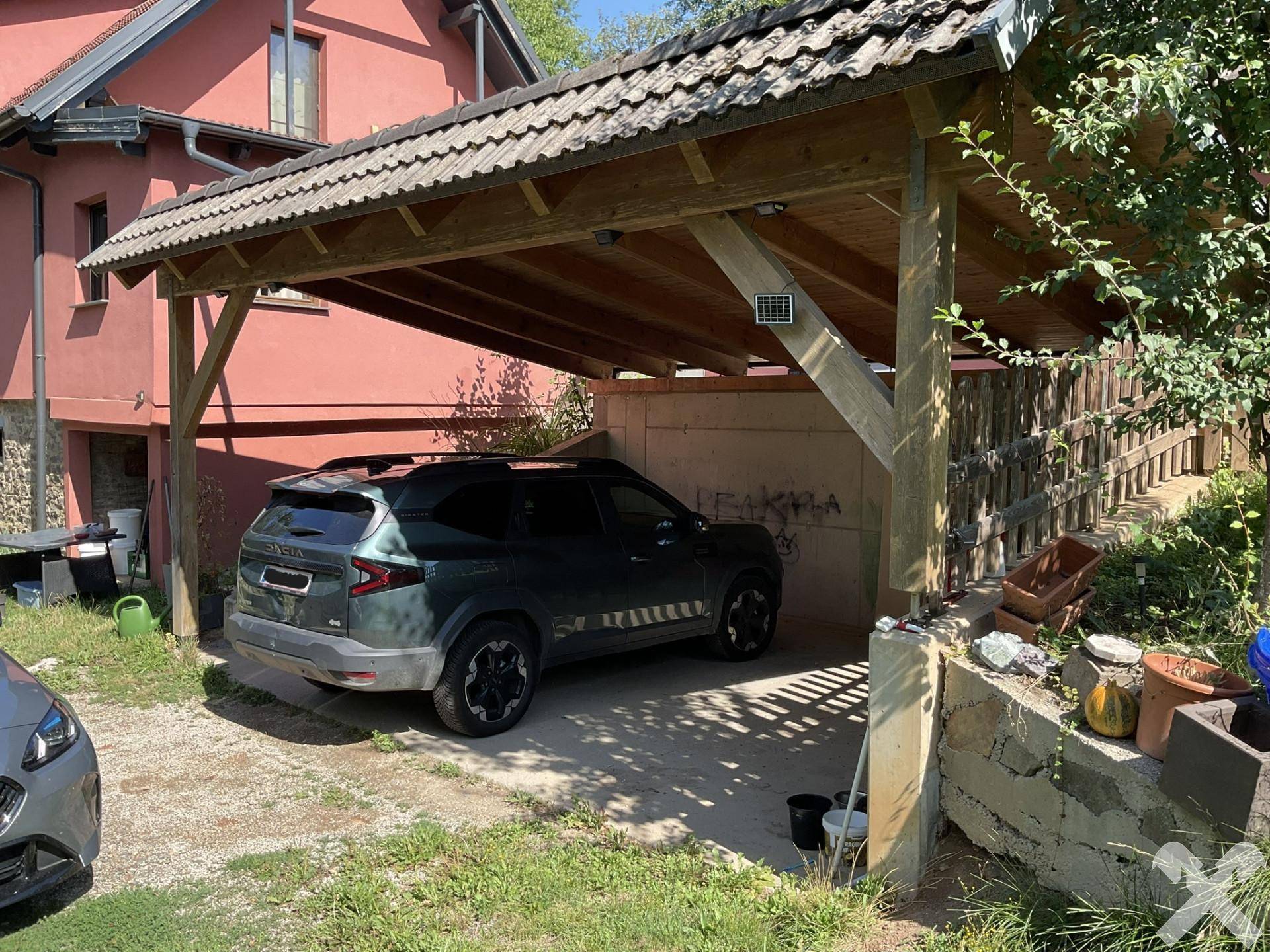 Überdachter Holzcarport mit einem geparkten Auto, vor der roten Fassade des Hauses gelegen.