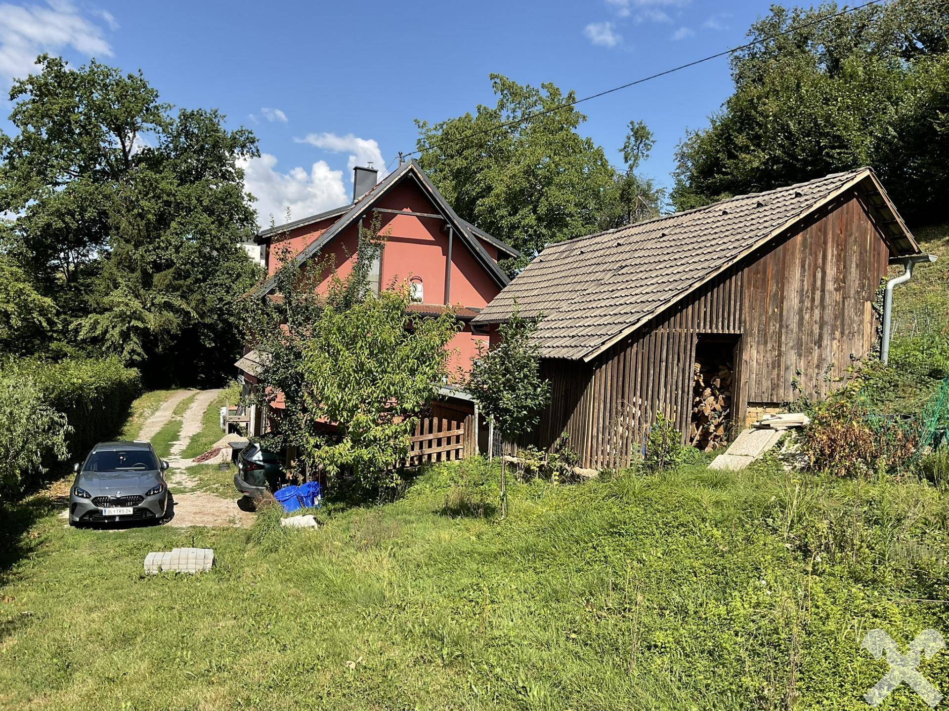 Rotes Haus mit Holzschuppen und Schotterauffahrt, umgeben von üppigem Grün und Bäumen.