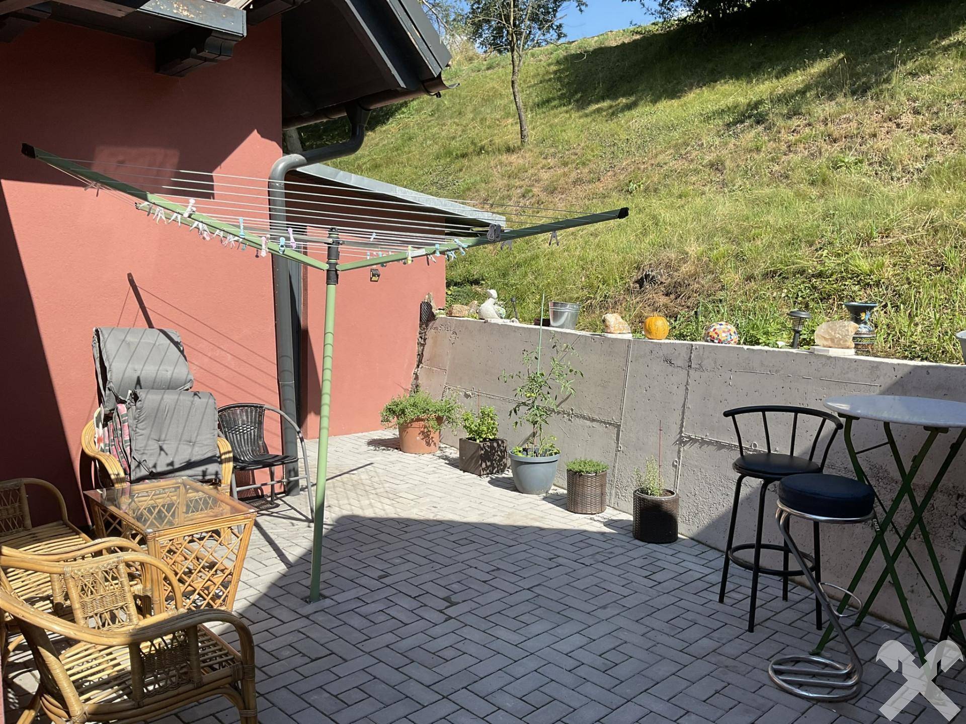 Gepflasterte Terrasse mit Korbmöbeln und Wäscheständer, umgeben von einer Betonmauer und Grün.