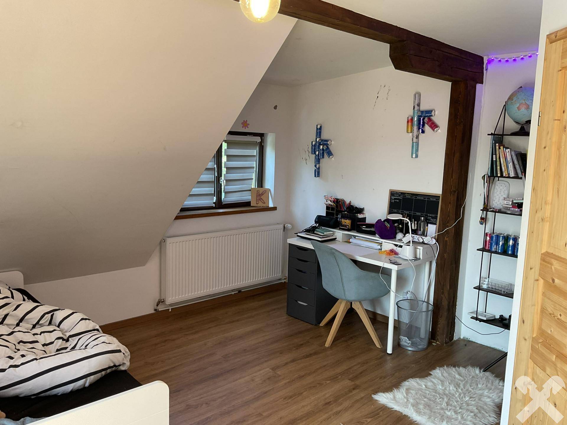 Kinderzimmer mit Schreibtisch, Regalen und sichtbaren Holzbalken, beleuchtet durch ein Fenster.