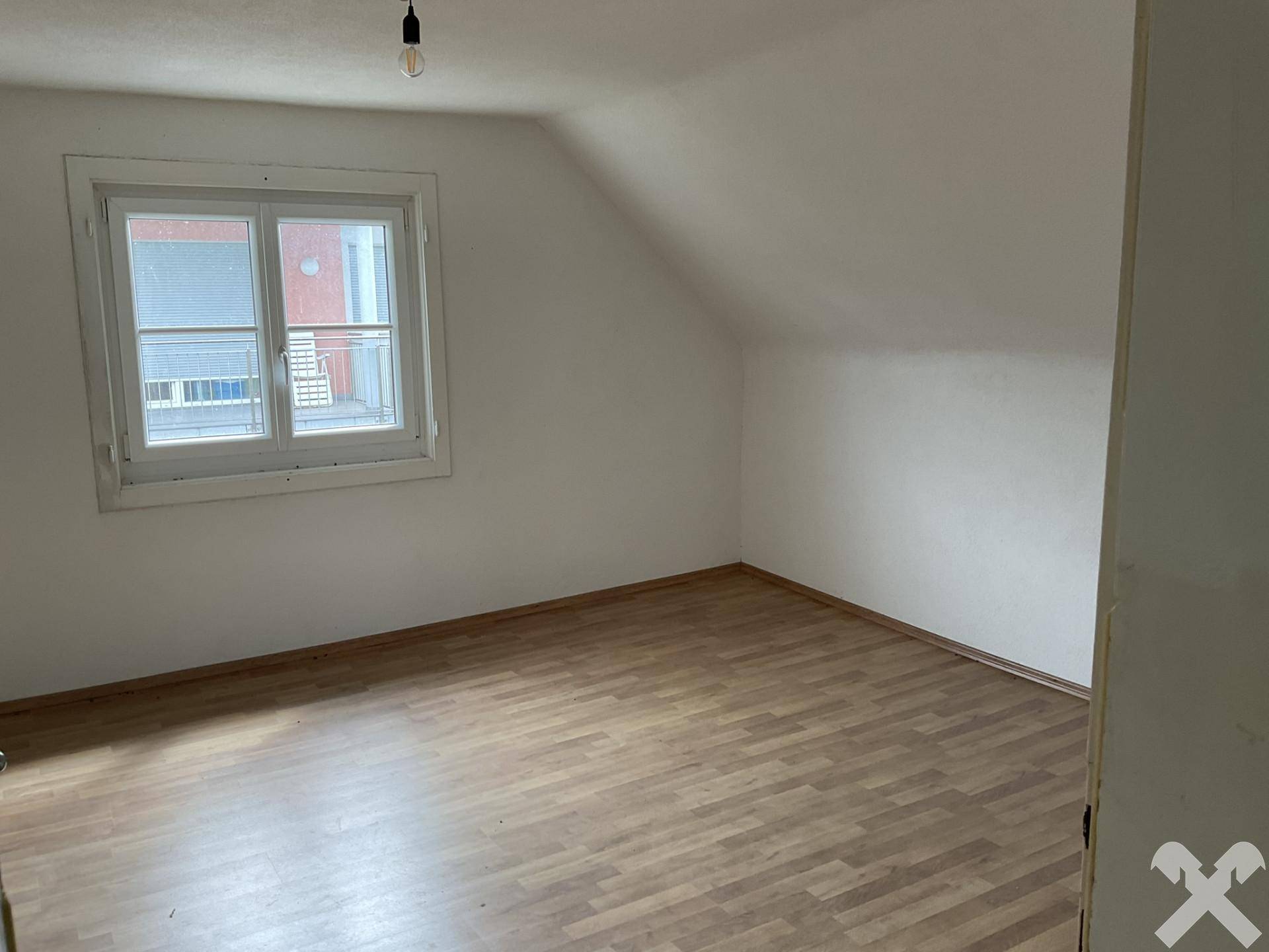 Helles Dachzimmer mit Laminatboden und einem Fenster, das den Blick auf die Nachbarschaft freigibt.
