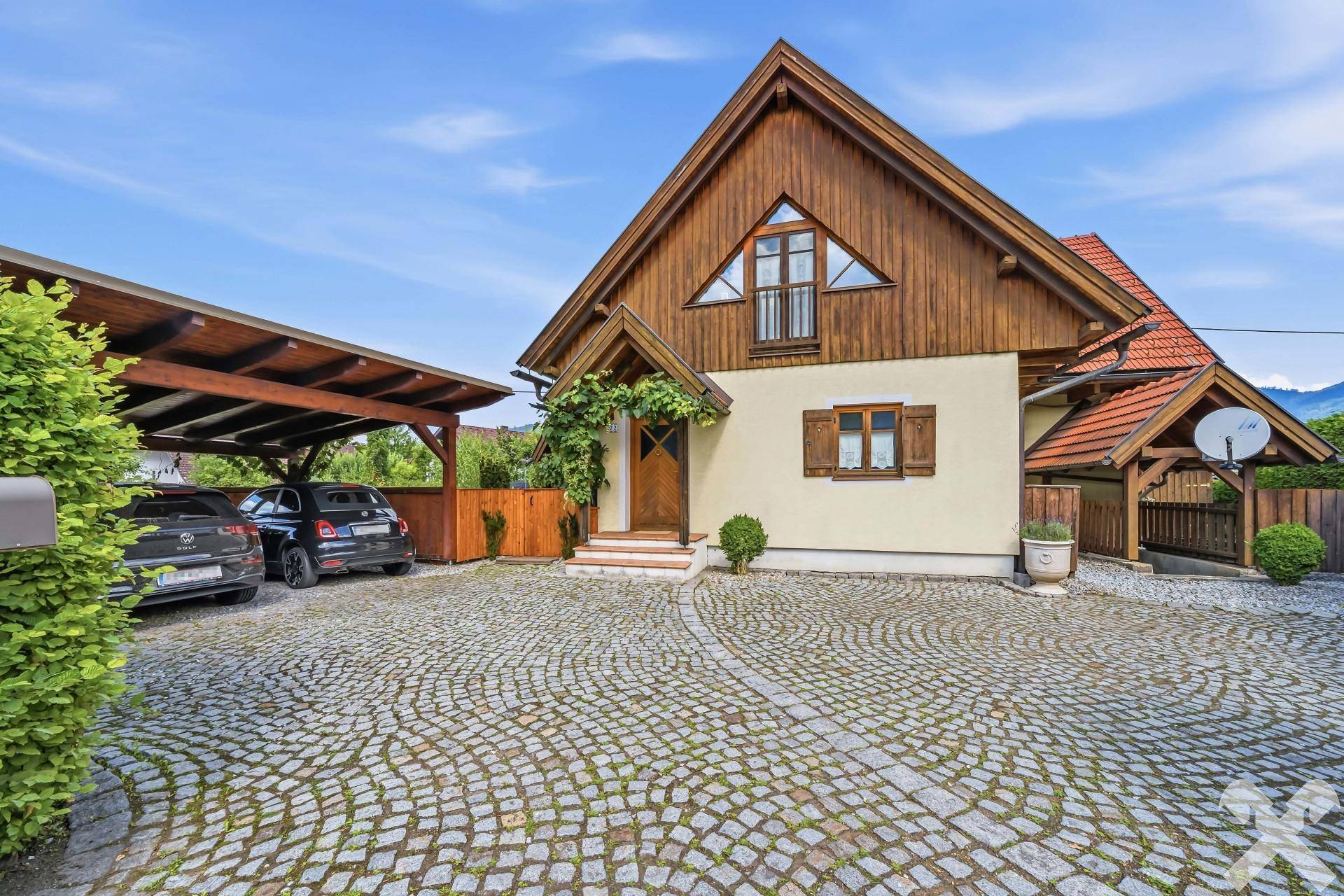 Charmantes Haus mit Holzfassade, gepflasterter Auffahrt und einem überdachten Carport für Fahrzeuge.