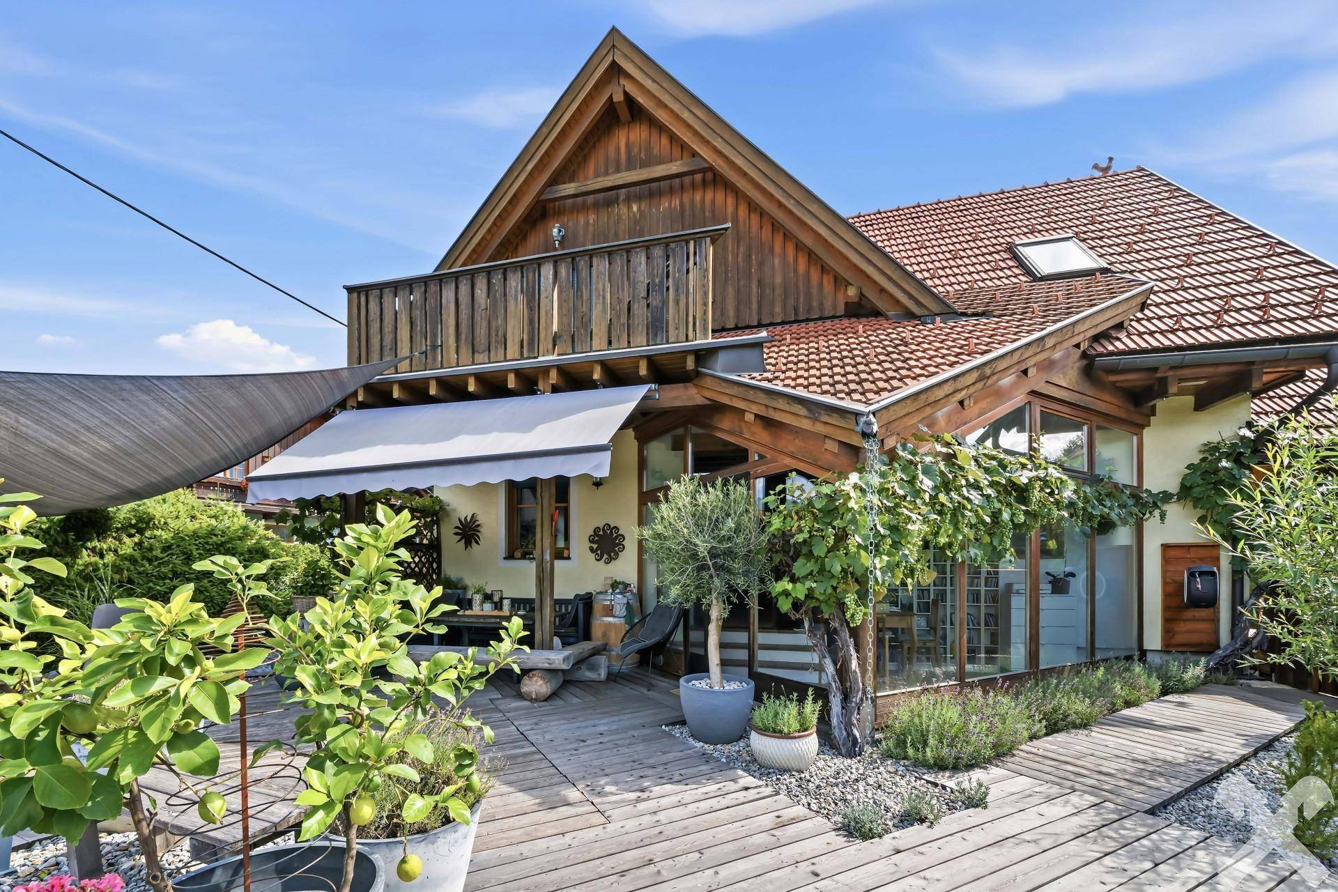 Große Holzterrasse mit Sonnenschutz, Pflanzen und Blick auf das charmante Haus mit Holzelementen.