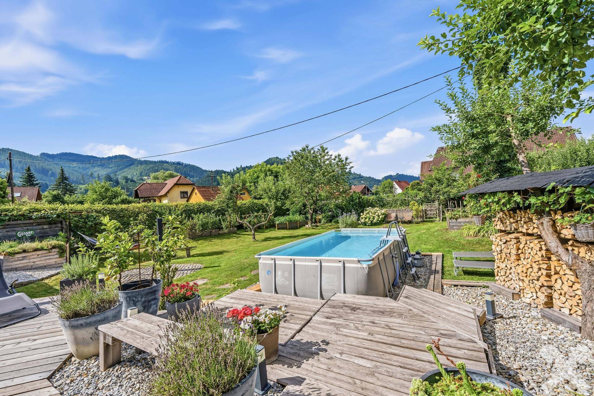 Holzterrasse mit Topfpflanzen und Blick auf den Aufstellpool sowie die umliegende grüne Landschaft.