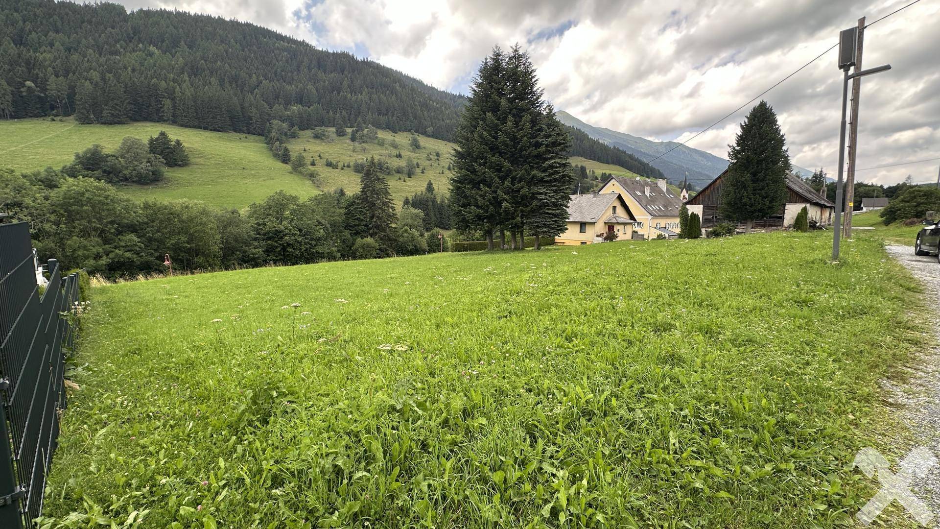 Grüne Wiese mit Blick auf die Berglandschaft und angrenzende Häuser unter bewölktem Himmel.