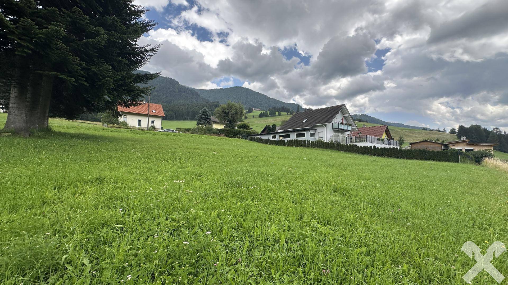 Idyllische Wiesenlandschaft mit vereinzelten Häusern und bewaldeten Berghängen im Hintergrund.