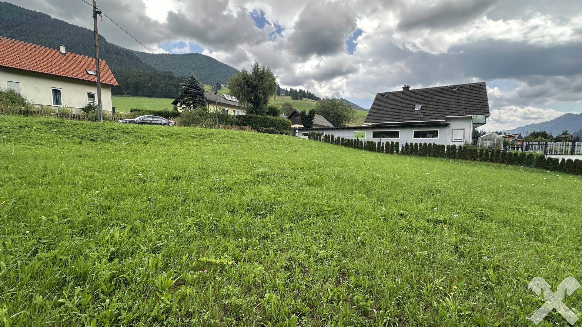 Grüne Wiese mit Blick auf die umliegende Bebauung und die Bergkulisse unter bewölktem Himmel.