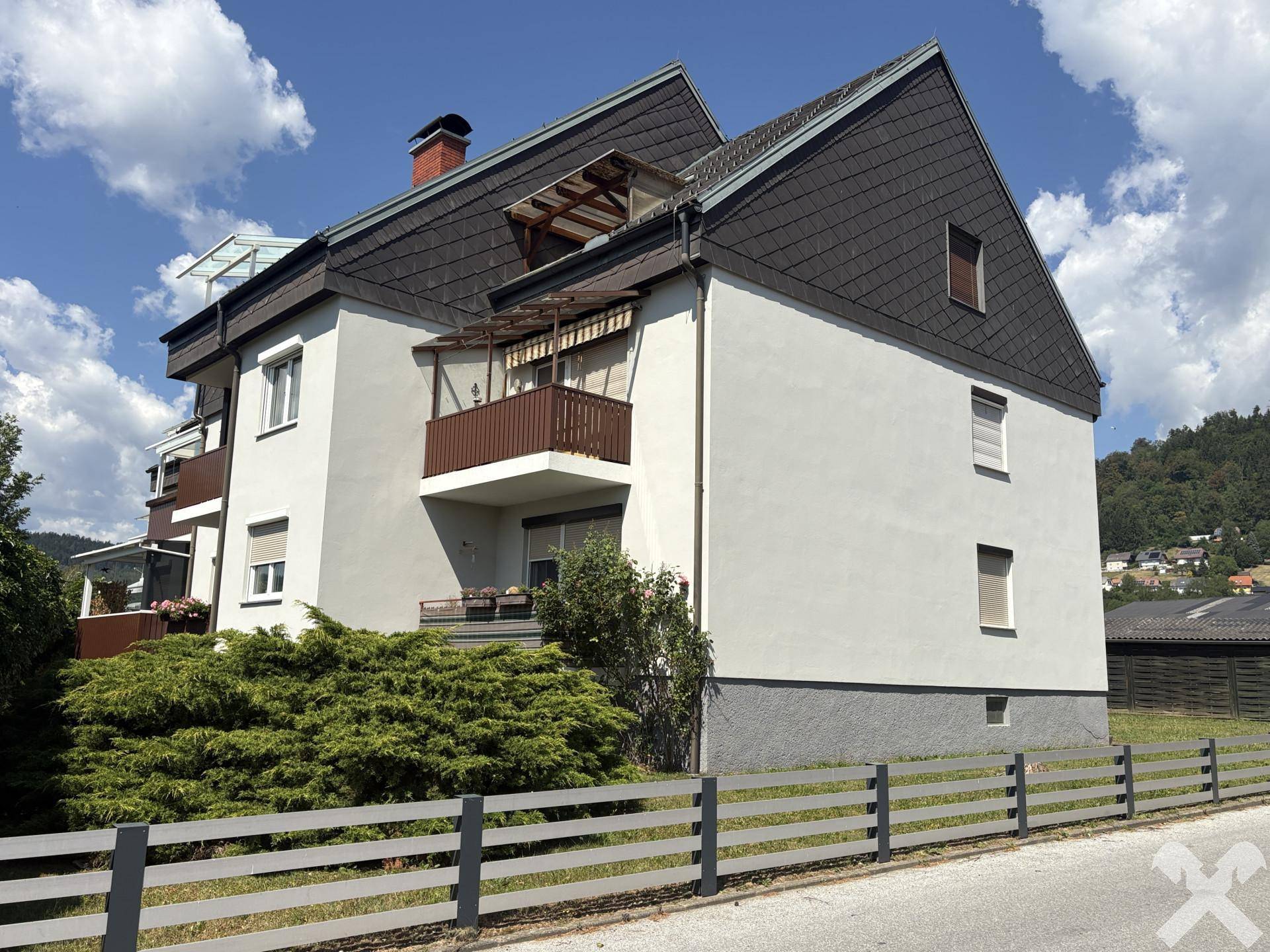 Seitenansicht des Wohngebäudes mit Satteldach, Balkonen und einem Zaun im Vordergrund unter blauem Himmel.