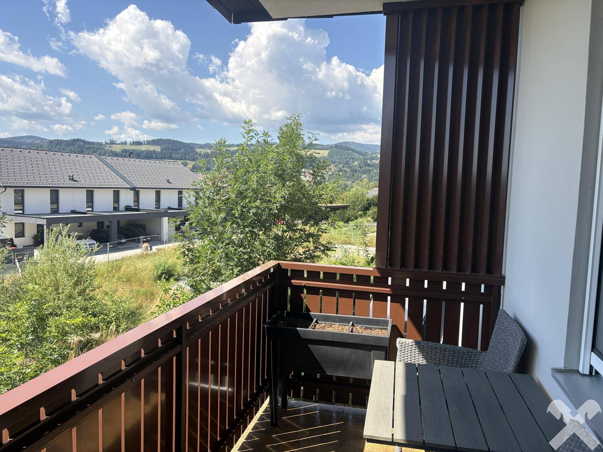 Möblierter Balkon mit Holzboden und weitem Blick über grüne Hügel und Nachbarhäuser an einem sonnigen Tag.