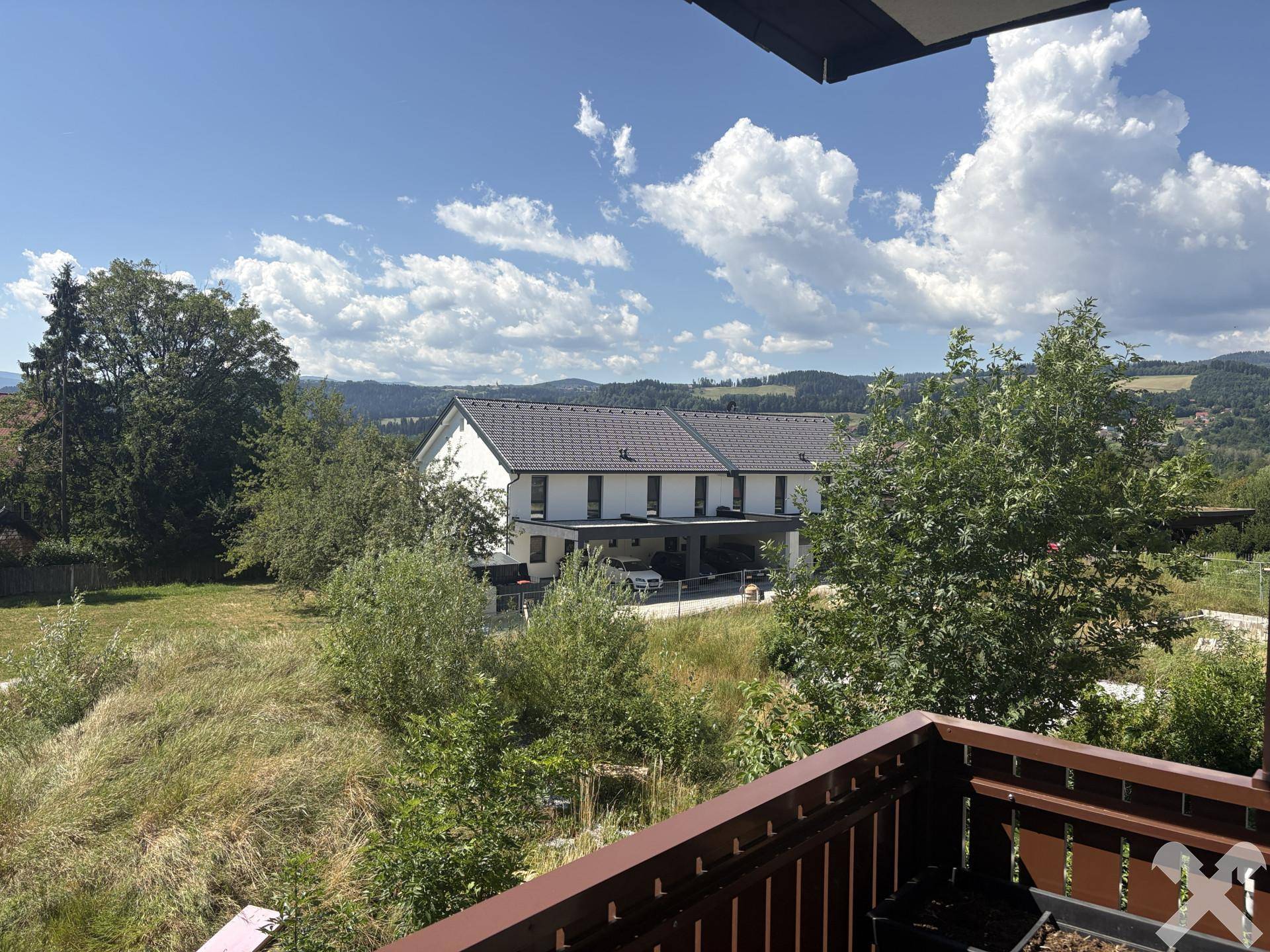 Weitblick vom Balkon über eine grüne Landschaft mit Bäumen und einem weiteren Wohngebäude unter blauem Himmel.