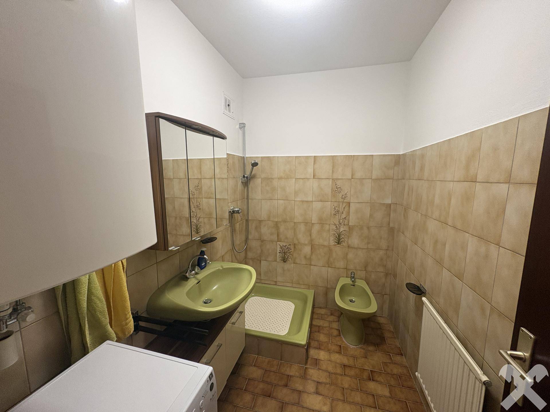 Badezimmer mit beigefarbenen Fliesen, Dusche, Waschbecken, Bidet und Heizkörper in 80er-Jahre-Stil.