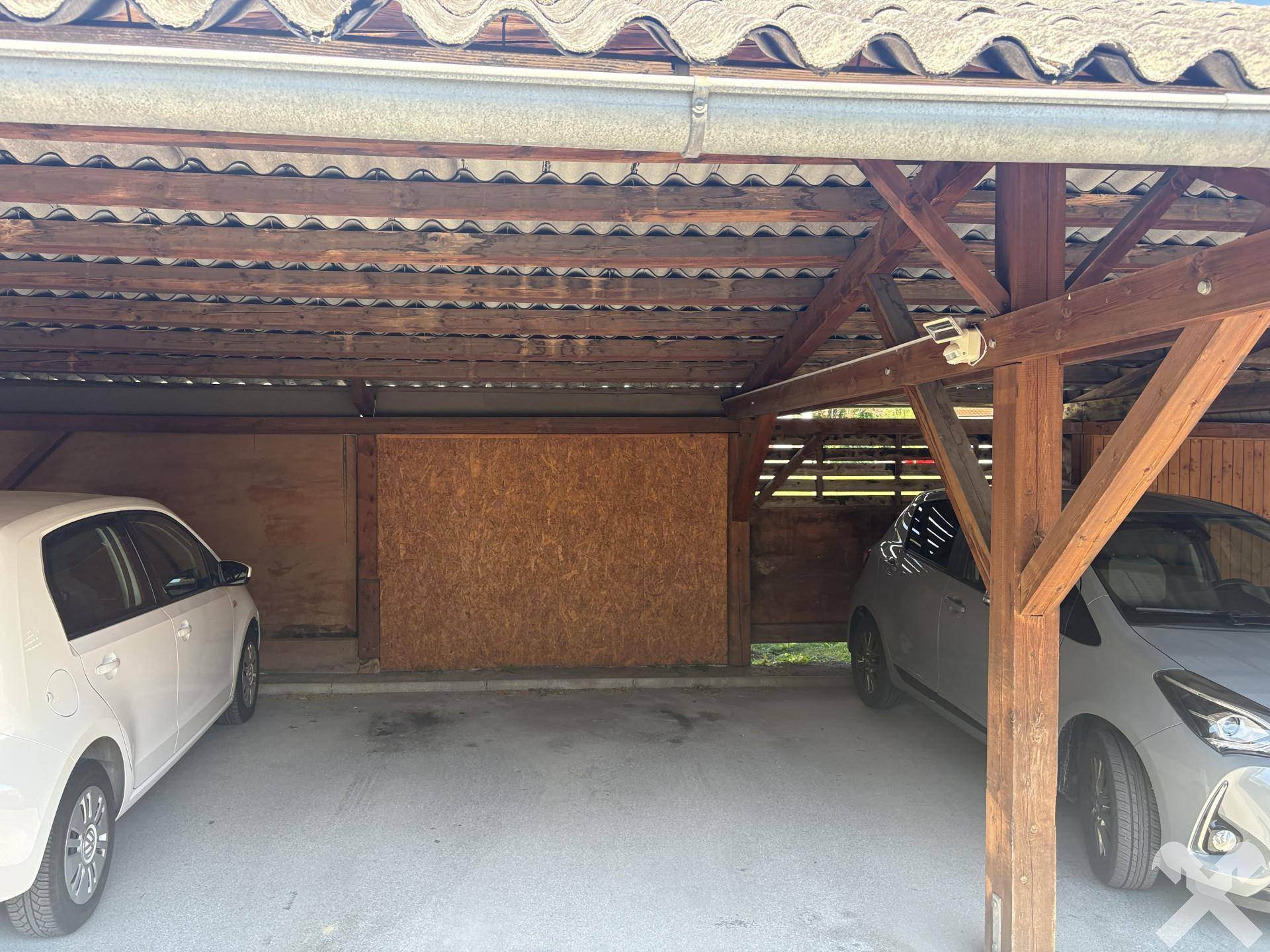 Überdachter Carport mit Holzbalken und zwei geparkten Autos an einem sonnigen Tag.