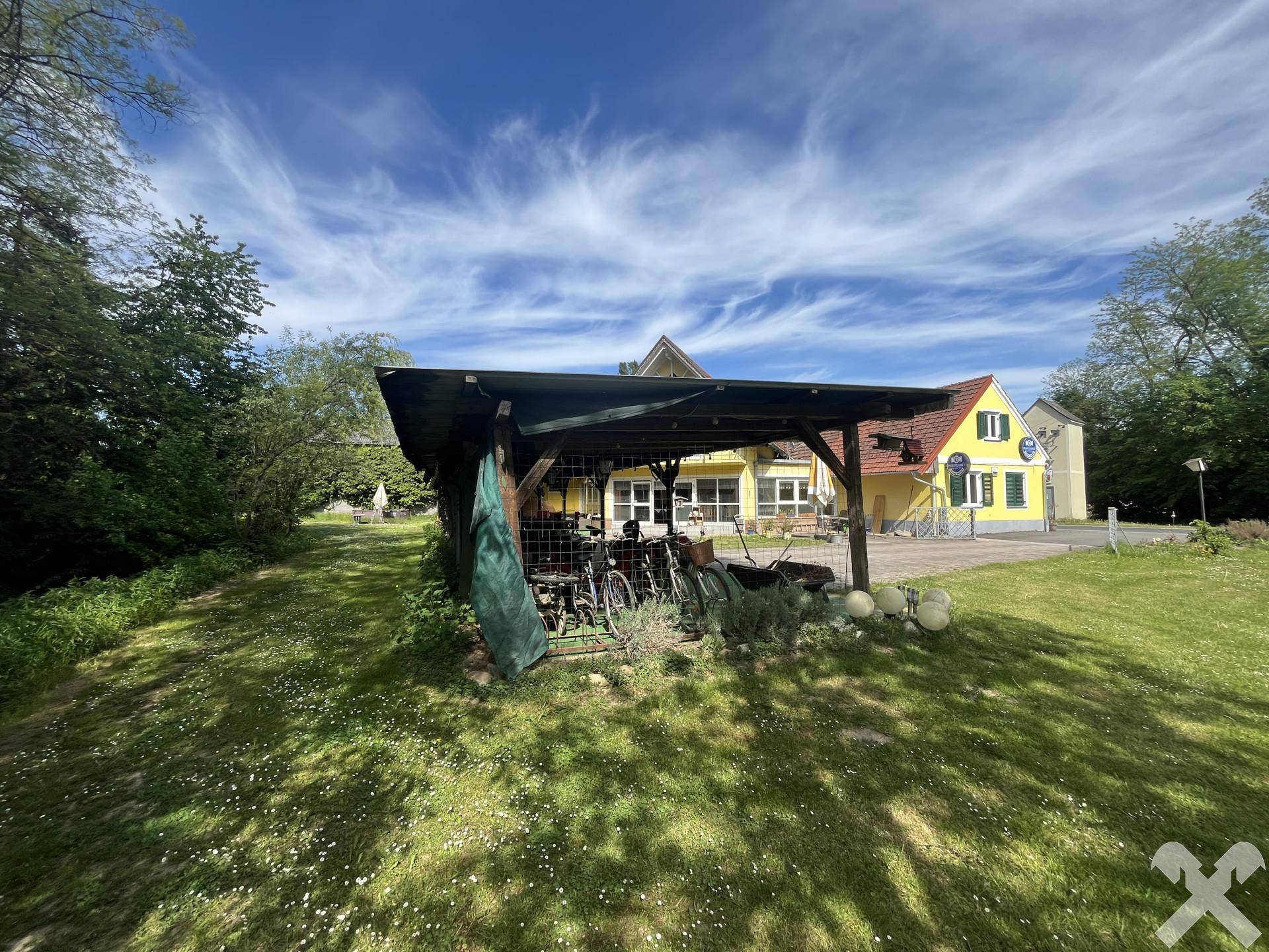 Holz-Carport auf einer grünen Wiese mit dem gelben Gebäude im Hintergrund unter blauem Himmel.