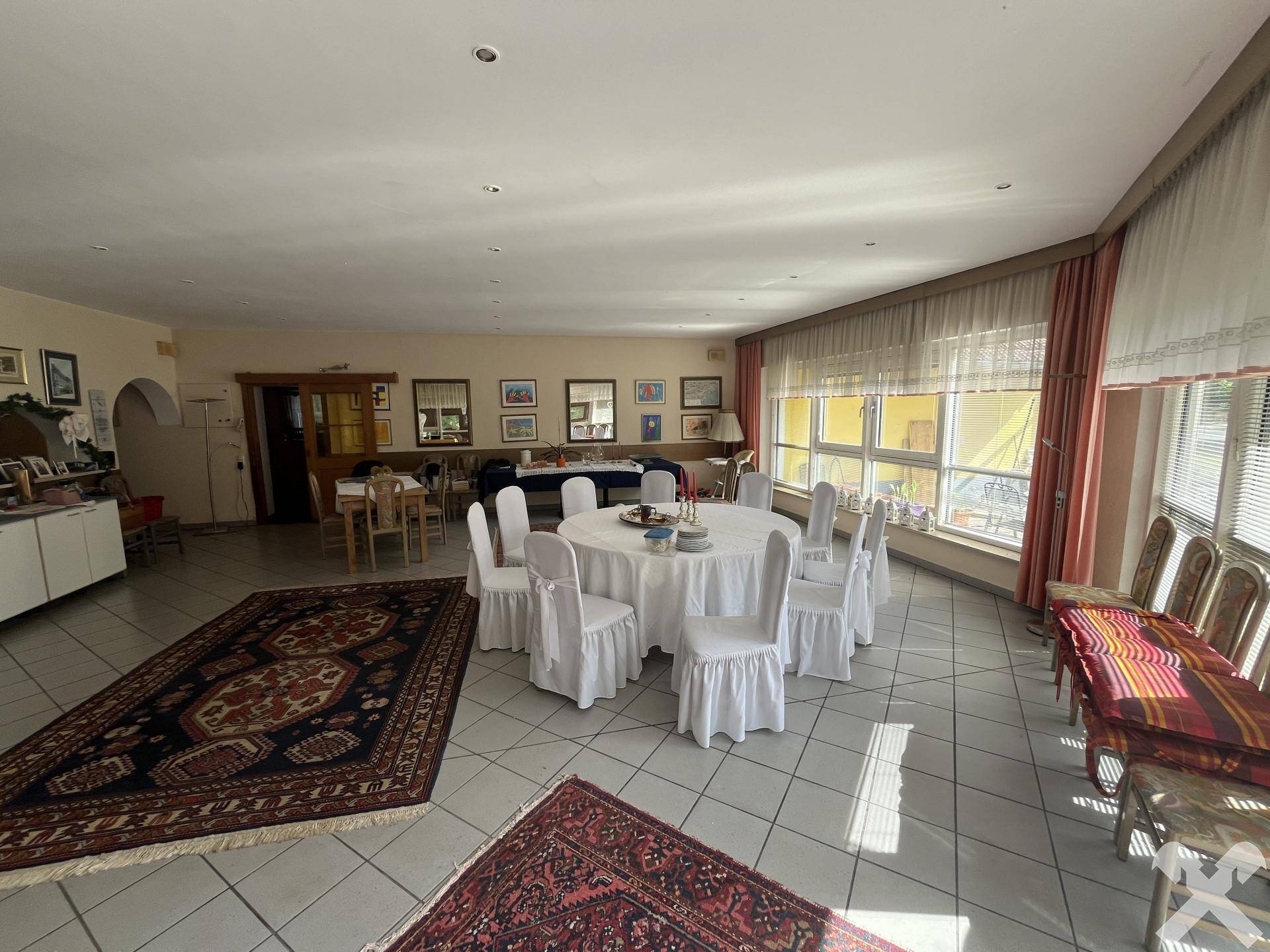 Großer Speisesaal mit vielen Tischen, Stühlen und Teppichen, hell durch große Fenster.