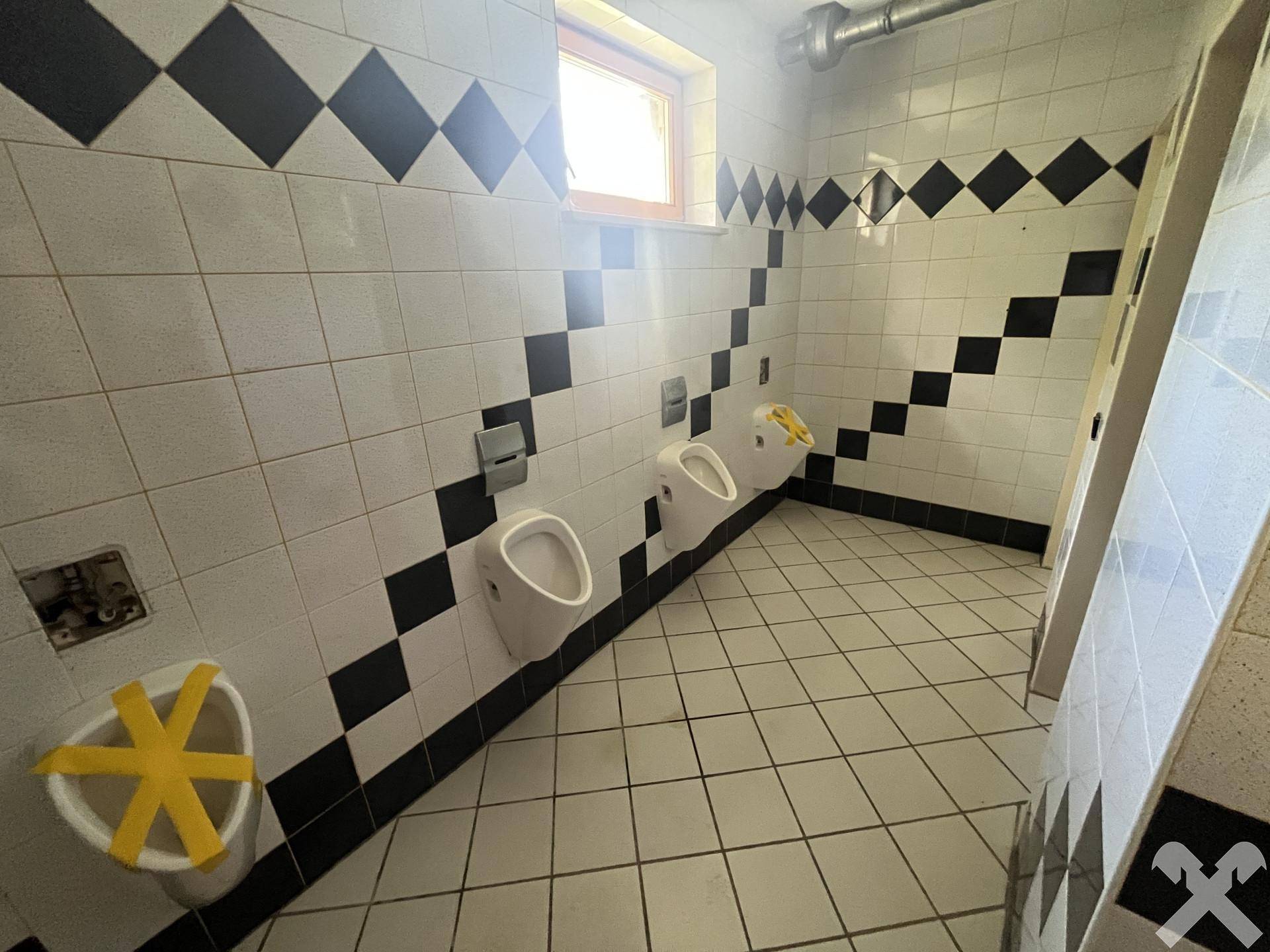 Herren-WC mit mehreren Urinalen, gefliesten Wänden in Schwarz-Weiß und einem Fenster.