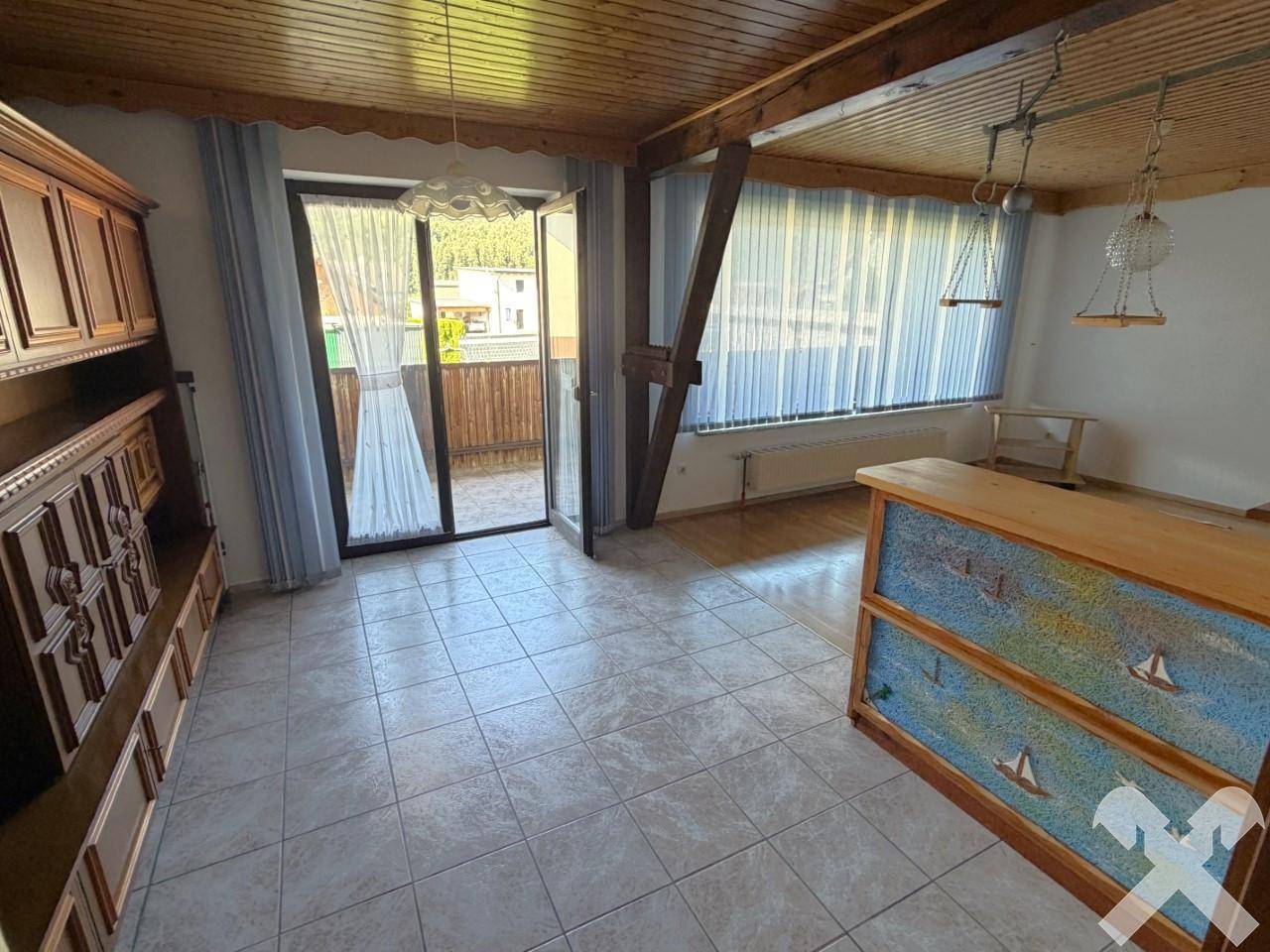 Geräumiger Wohnbereich mit Fliesenboden, Holzbalken und Zugang zum Balkon.