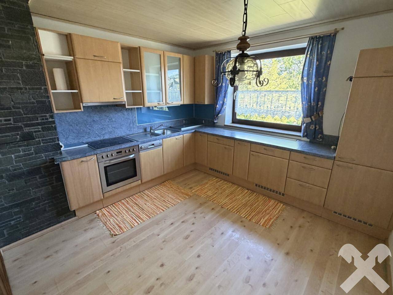 Funktionale Küche mit Holzschränken, blauer Arbeitsplatte und Fenster mit Blick ins Grüne.