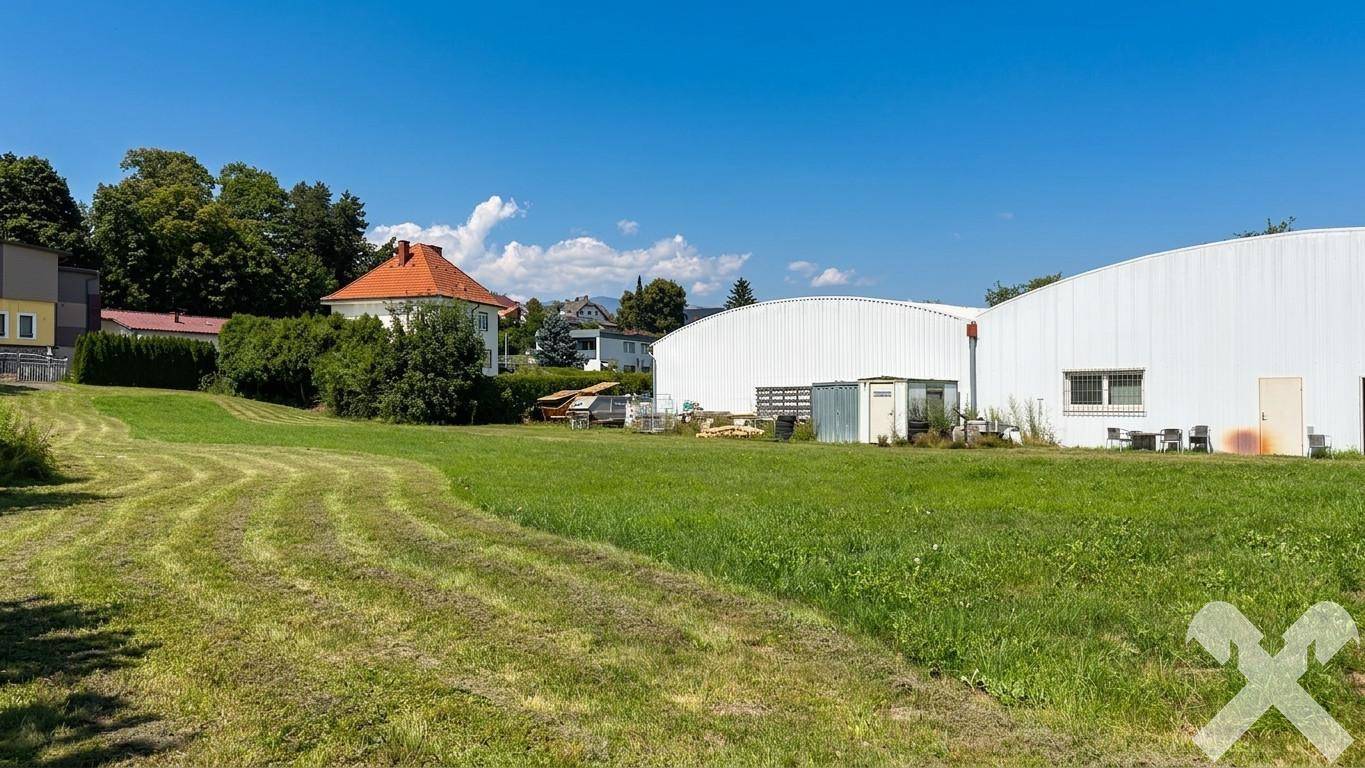 Weitläufige grüne Wiese mit angrenzenden Industriegebäuden und einem traditionellen Wohnhaus im Hintergrund.