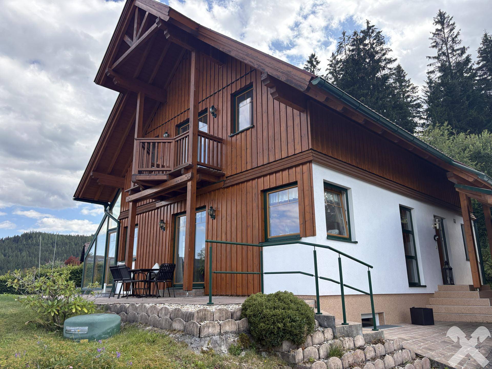 Ansicht des Hauses mit Holzverkleidung, kleinem Balkon und einer gepflasterten Terrasse im Grünen.