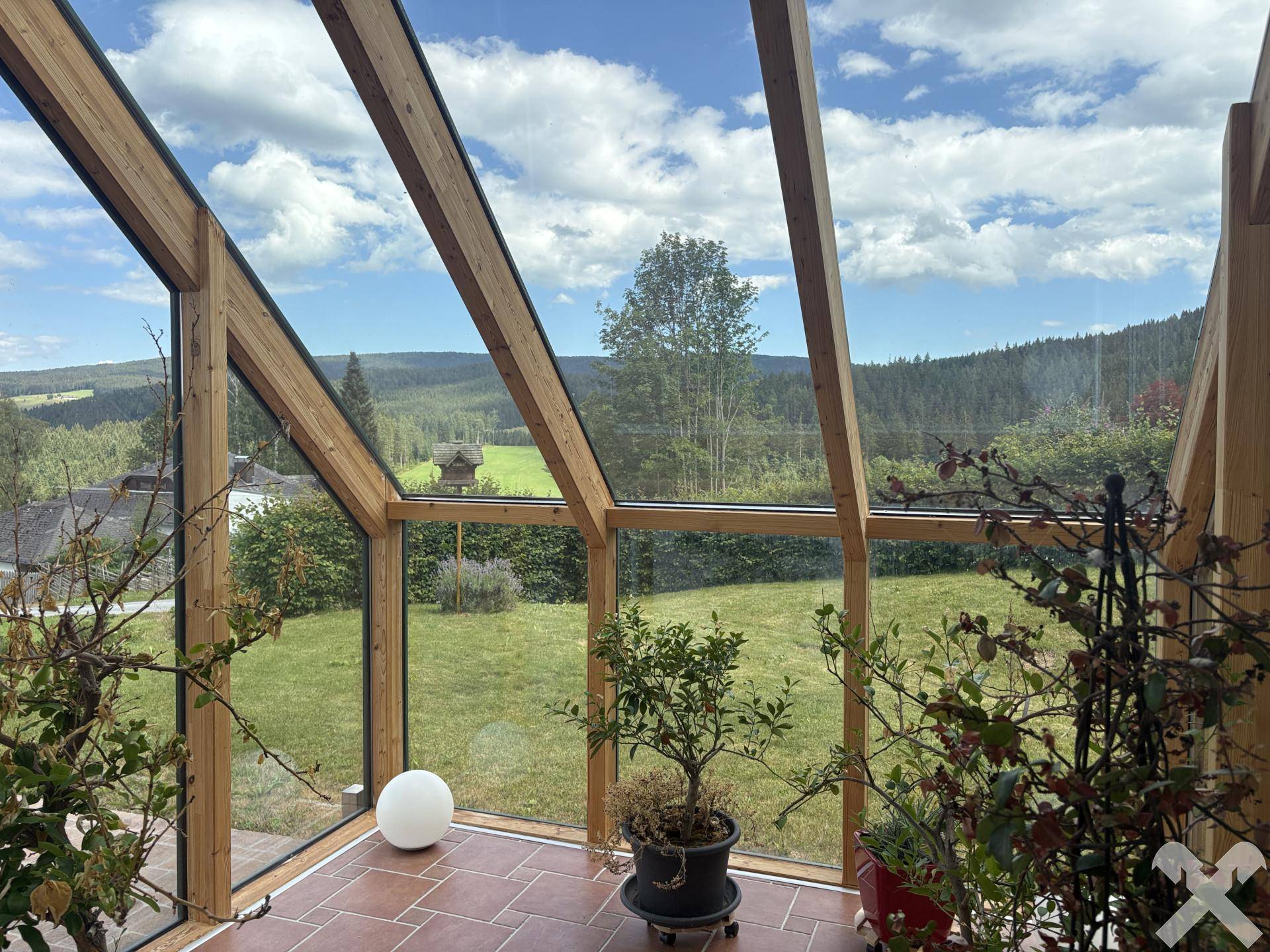 Heller Wintergarten mit Holzbalken, vielen Pflanzen und Panoramablick auf die grüne Landschaft.