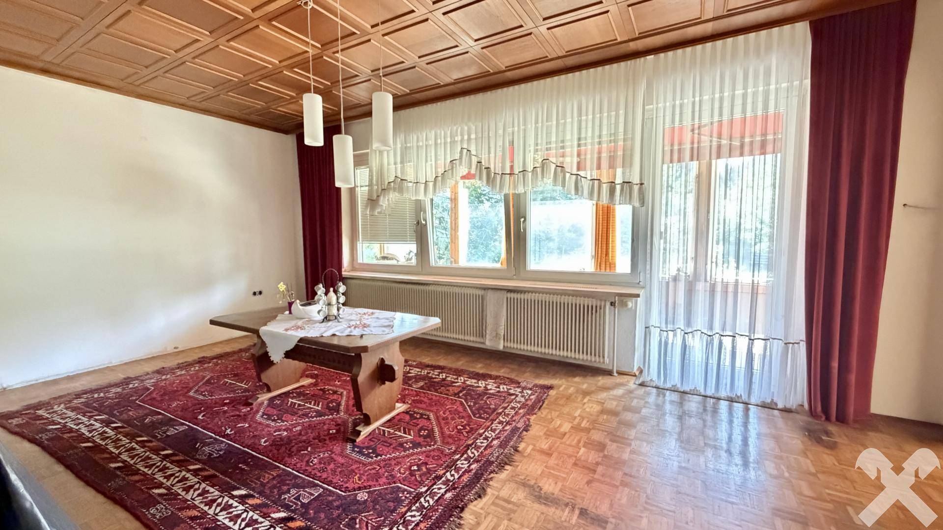 Geräumiges Wohnzimmer mit Holzboden, Kassettendecke und großen Fenstern mit Vorhängen.