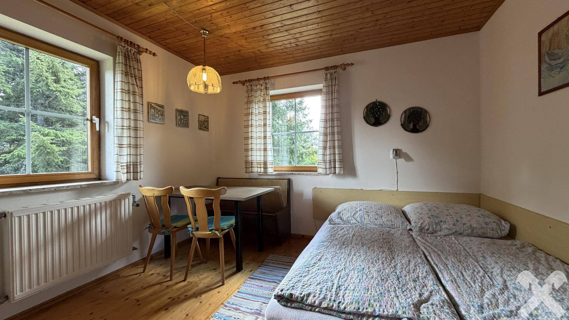Schlafzimmer mit Holzbalkendecke, Einzelbett und kleinem Essbereich mit Waldblick.