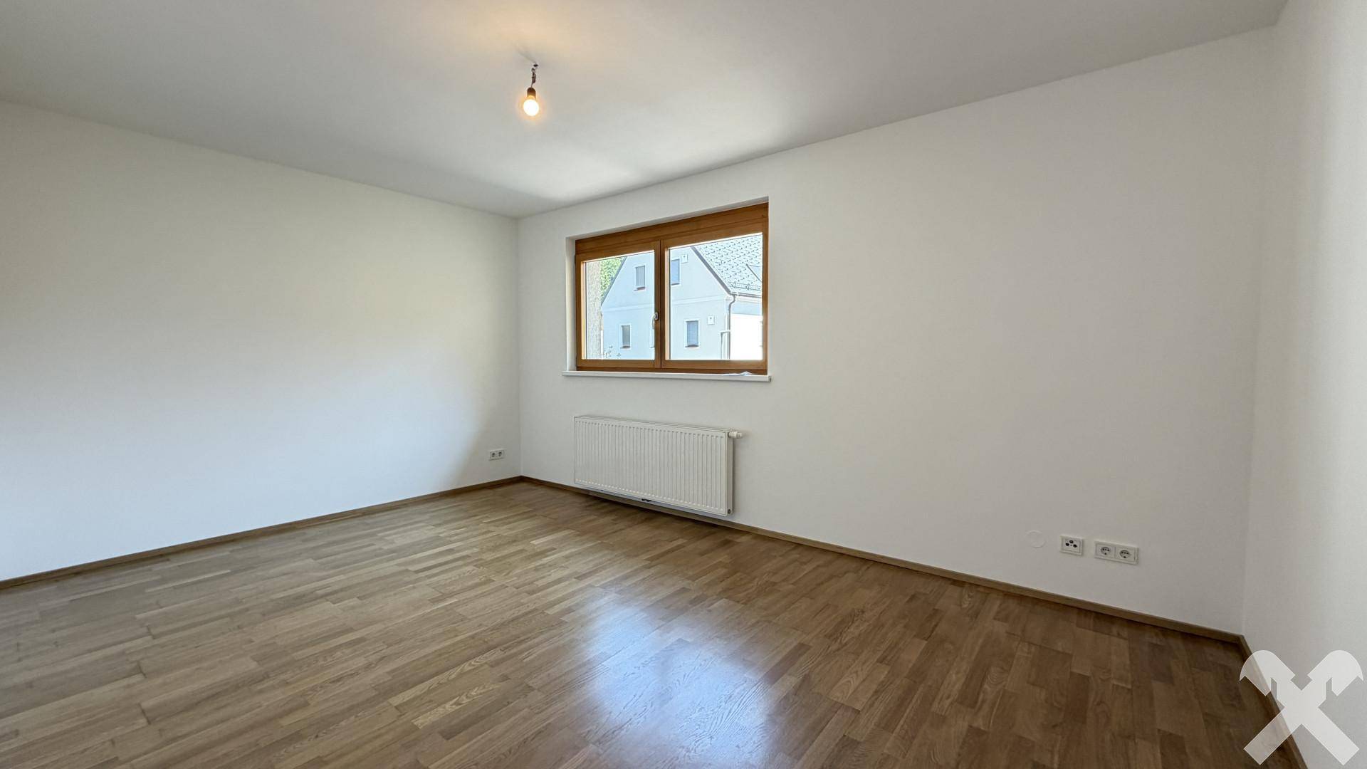 Helles, leeres Schlafzimmer mit Holzparkettboden und einem Fenster mit Blick auf die Nachbarschaft.