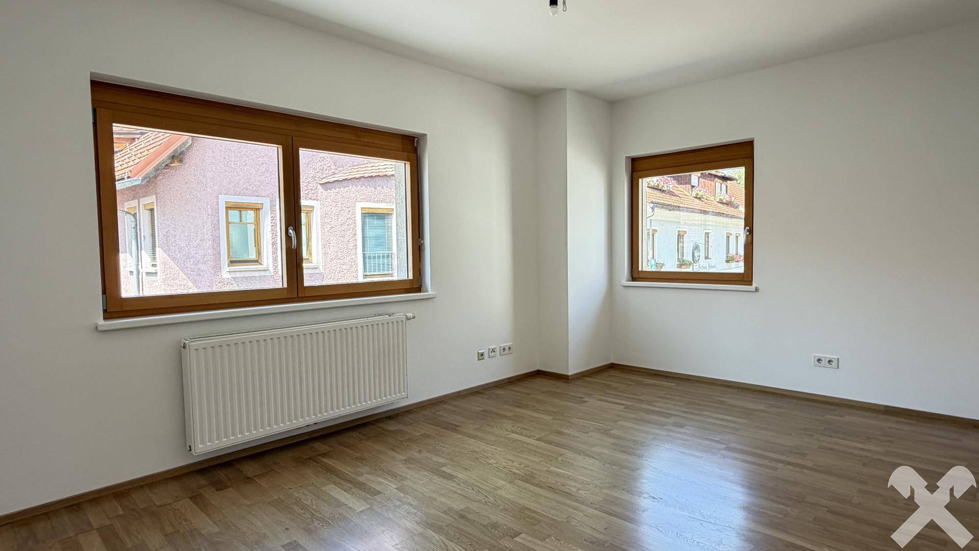 Leeres Schlafzimmer mit Holzparkettboden und zwei Fenstern, die viel Licht hereinlassen.